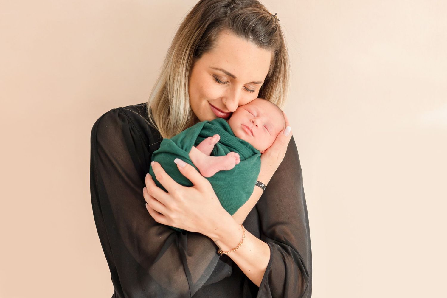 Sweet embrace with sleeping newborn wrapped in green against beige wall.