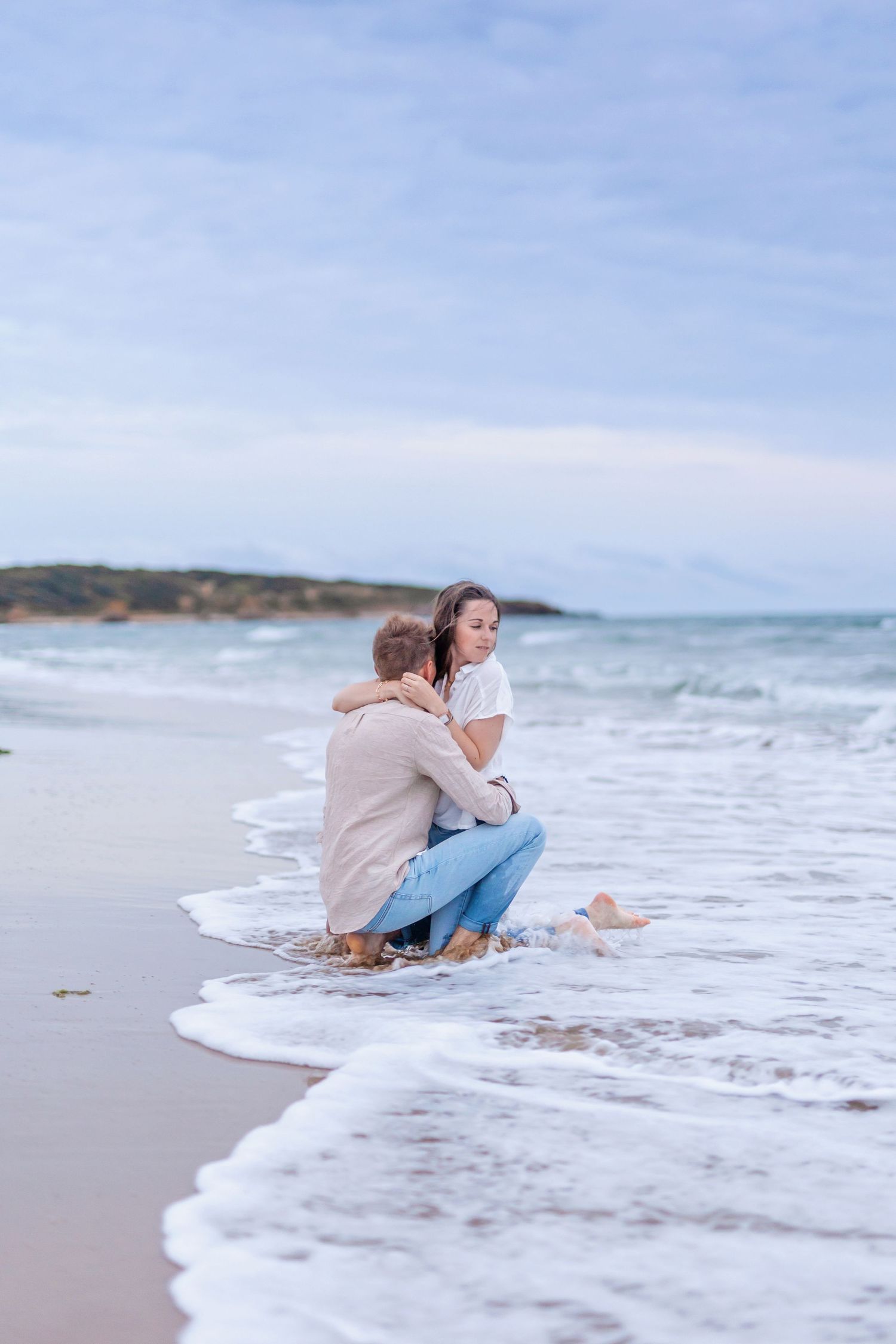 A couple sits together on the beach as waves wash around them during sunset.