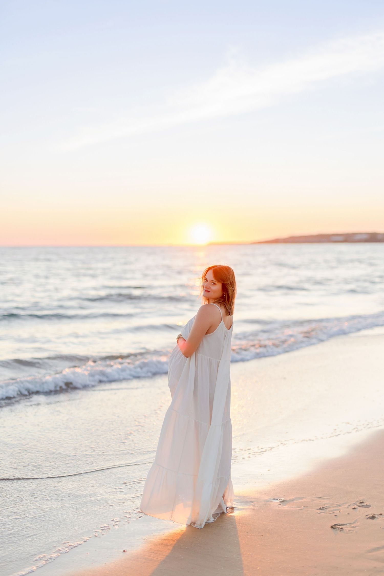 Person in white maxi dress stands on beach during golden hour sunset.