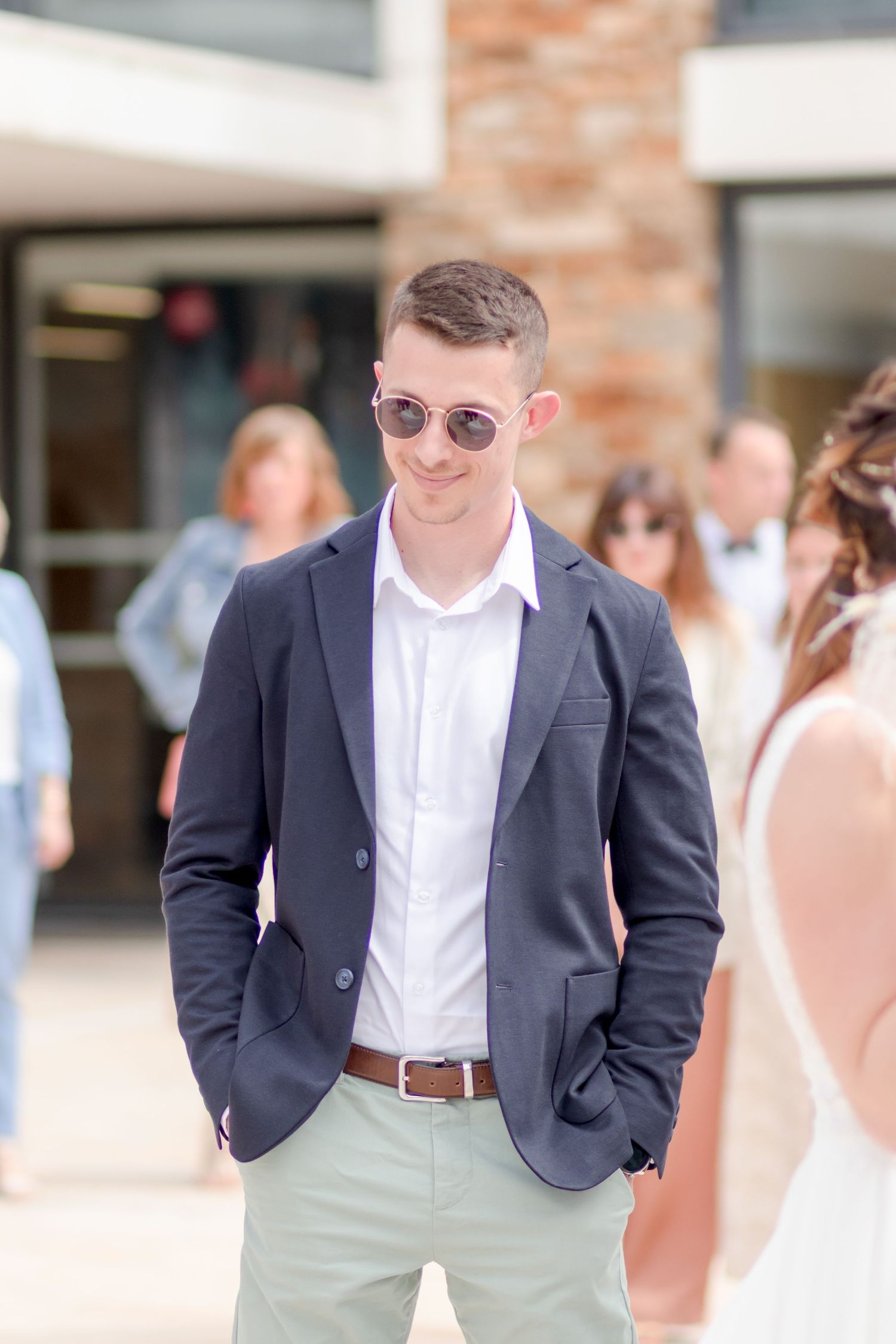 Wedding guest in navy blazer and white shirt poses outdoors near brick building.
