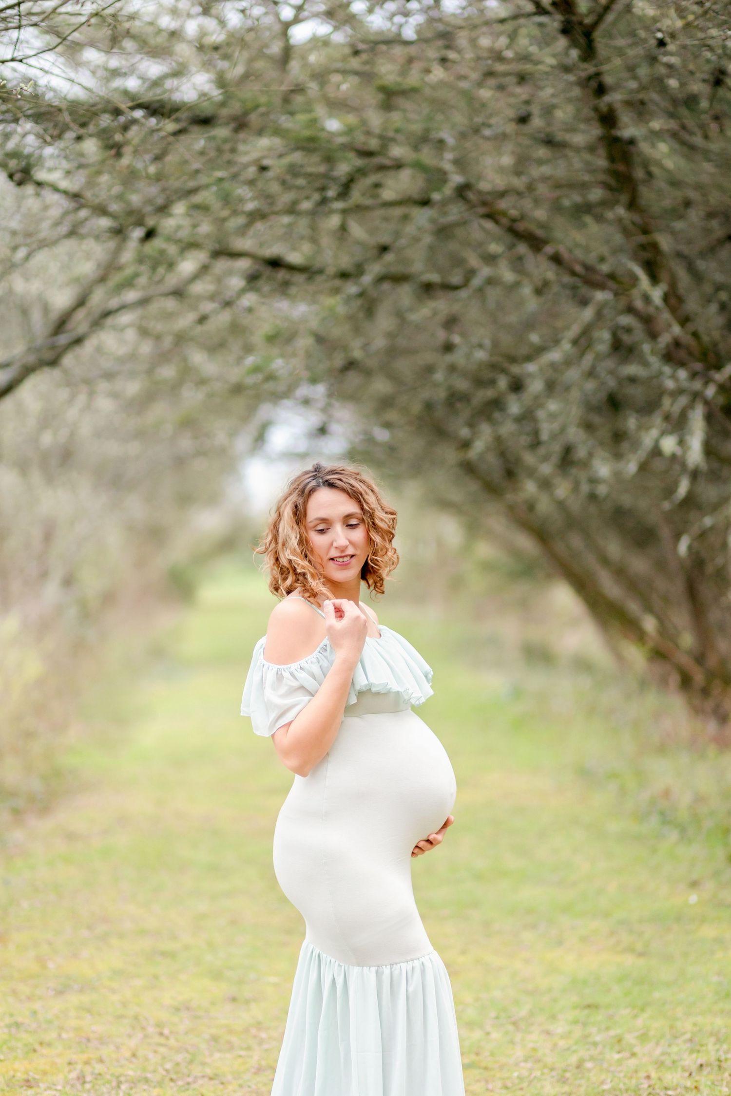 An ethereal maternity photo of a woman in a flowing white dress posing outdoors under trees.