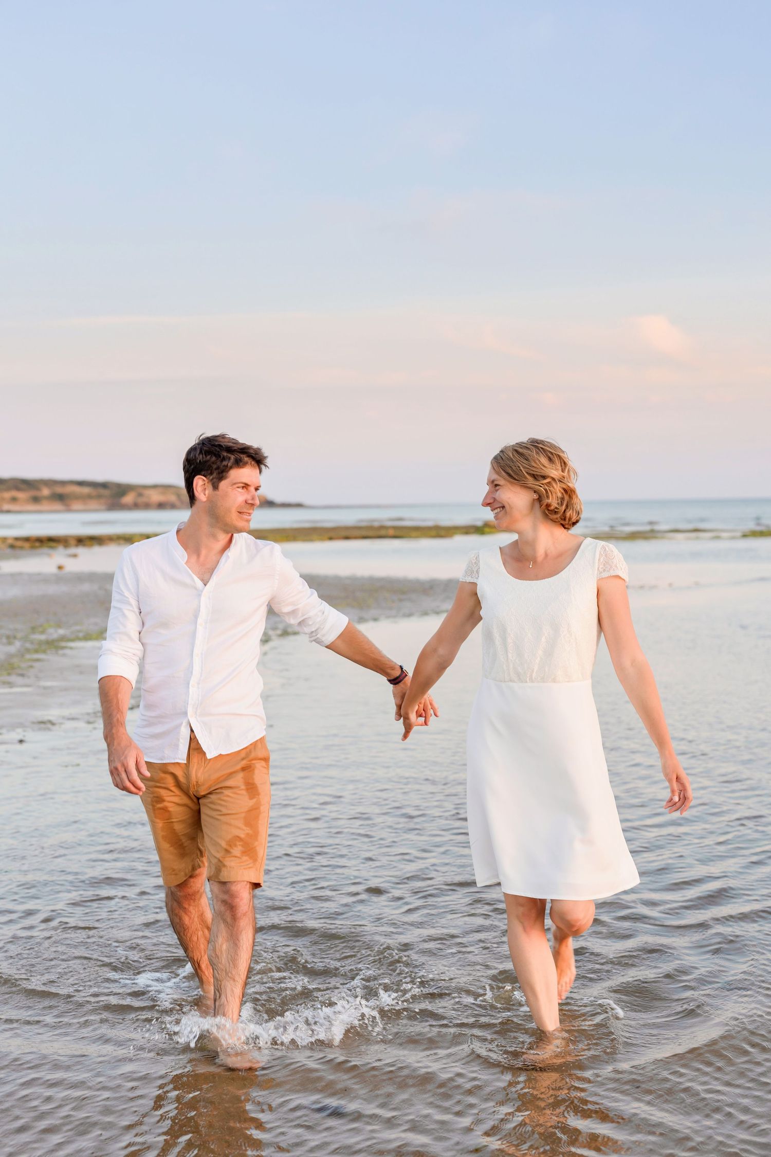 A playful couple splashes through shallow beach water wearing white and tan summer attire.