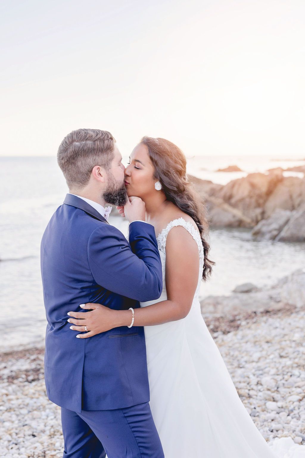 An intimate wedding kiss captured on a scenic beach with rocks and gentle waves.