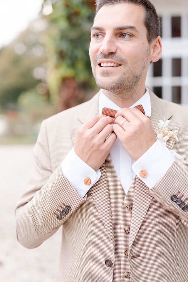 Groom in beige textured suit adjusts coral bowtie while smiling with boutonniere pinned to lapel.