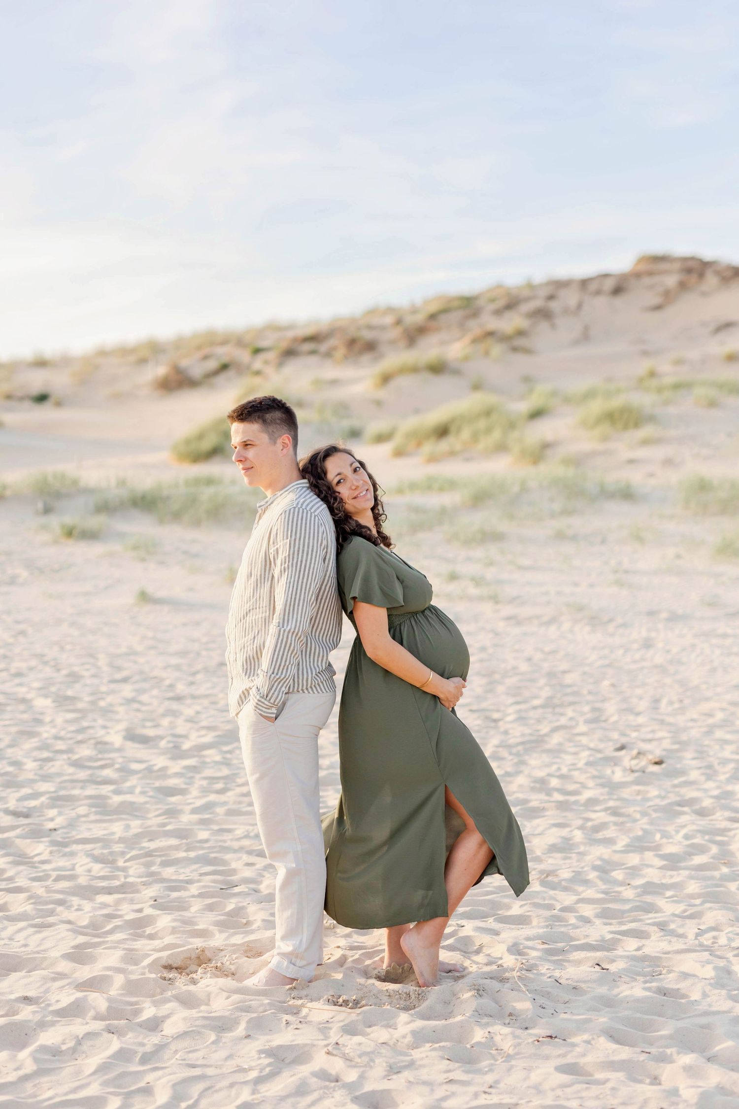 A couple poses back-to-back on sandy beach during maternity photoshoot.