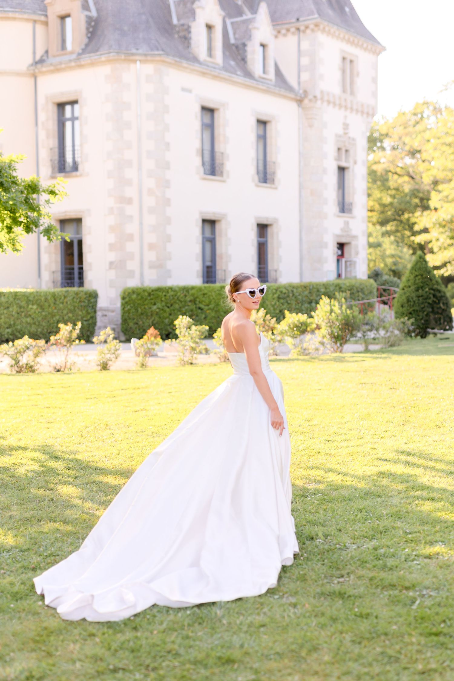 Wedding dress flowing on grass with elegant chateau backdrop in sunlight.