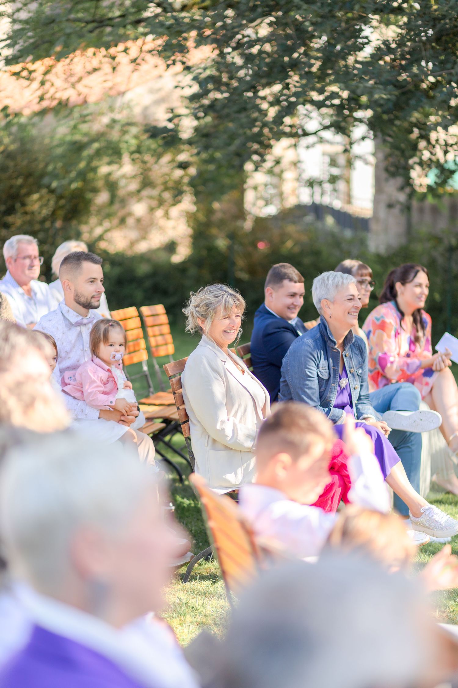 Wedding guests seated on wooden benches during outdoor ceremony under trees.