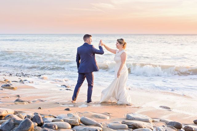 A couple in wedding attire dance together on a beach with waves and rocks at sunset.