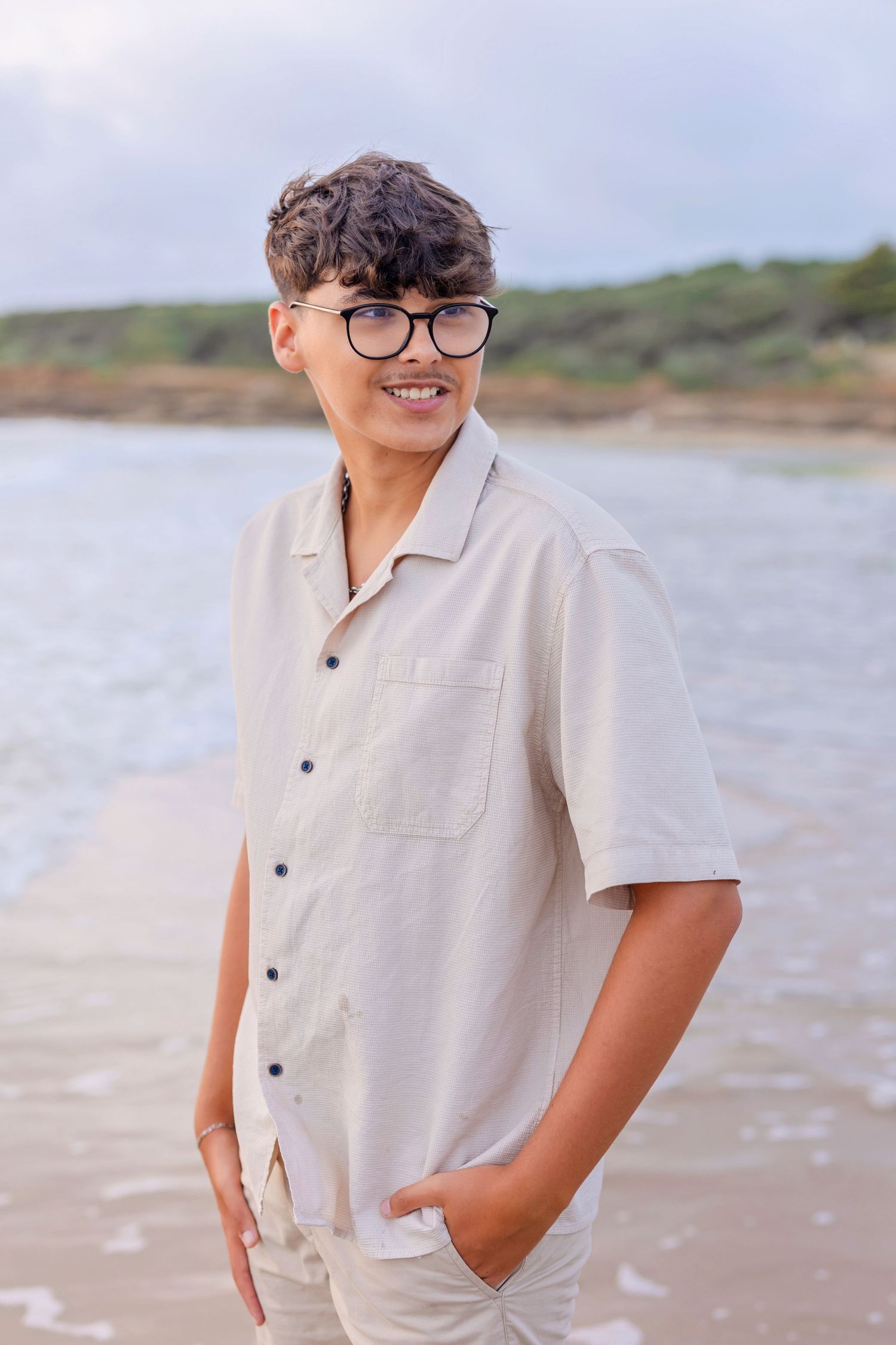 A person in glasses and white shirt smiling on a beach with water in background.