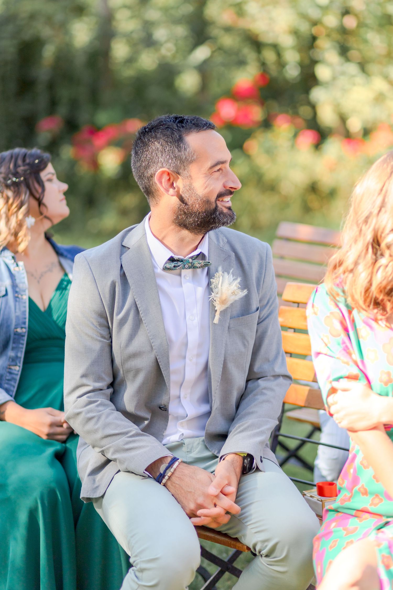 Wedding guests in formal attire seated on wooden benches at outdoor ceremony.