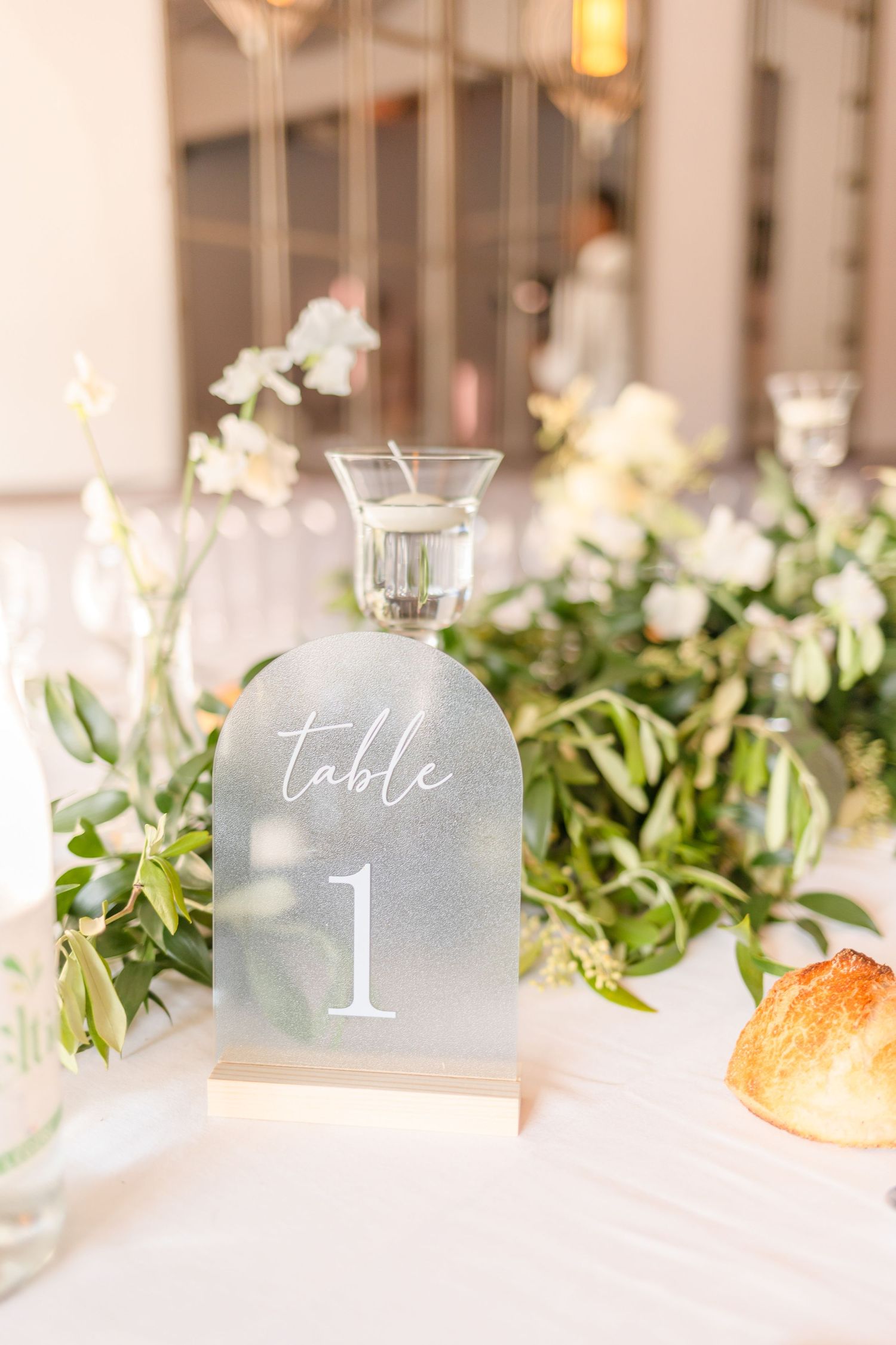 Frosted glass table number with greenery and white floral centerpiece at wedding reception.