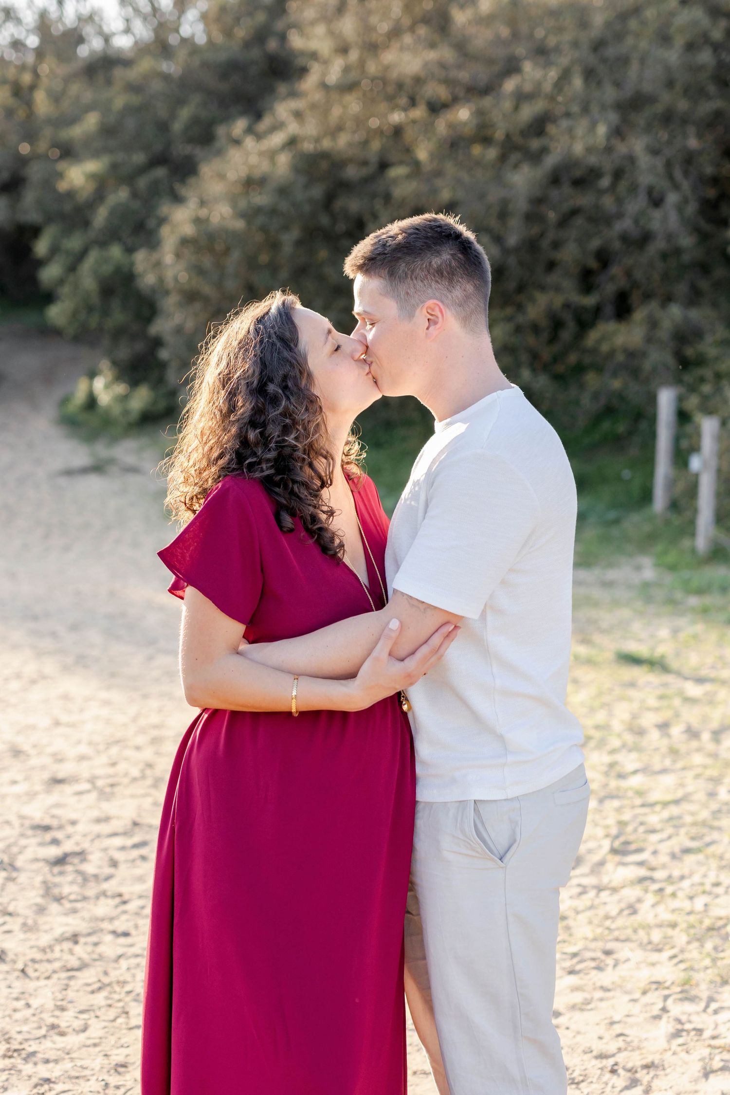A romantic couple shares a tender moment on a sandy beach path, with her wearing a flowing red dress.