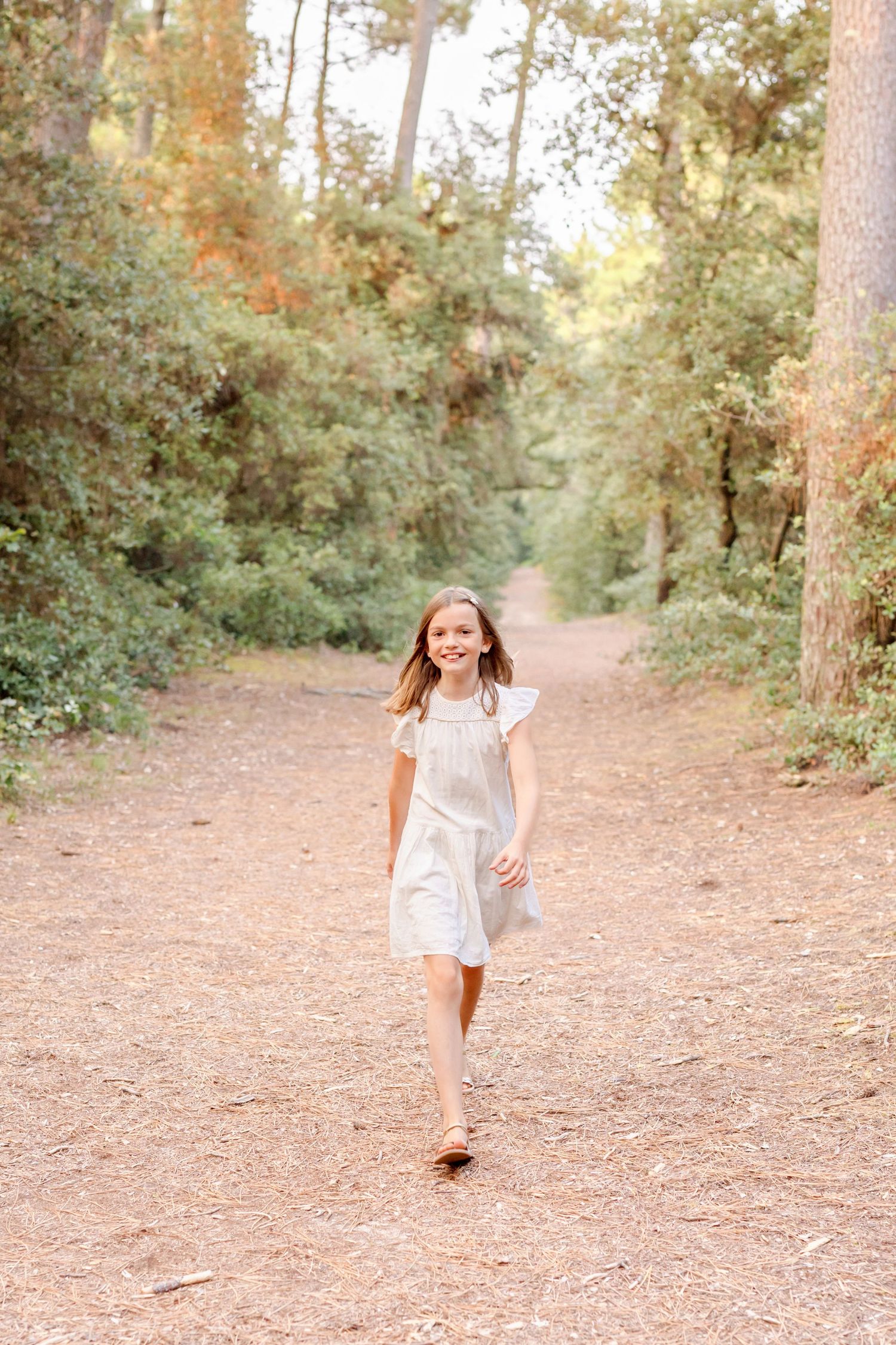 Young person in white dress walking cheerfully down a sunlit forest path.