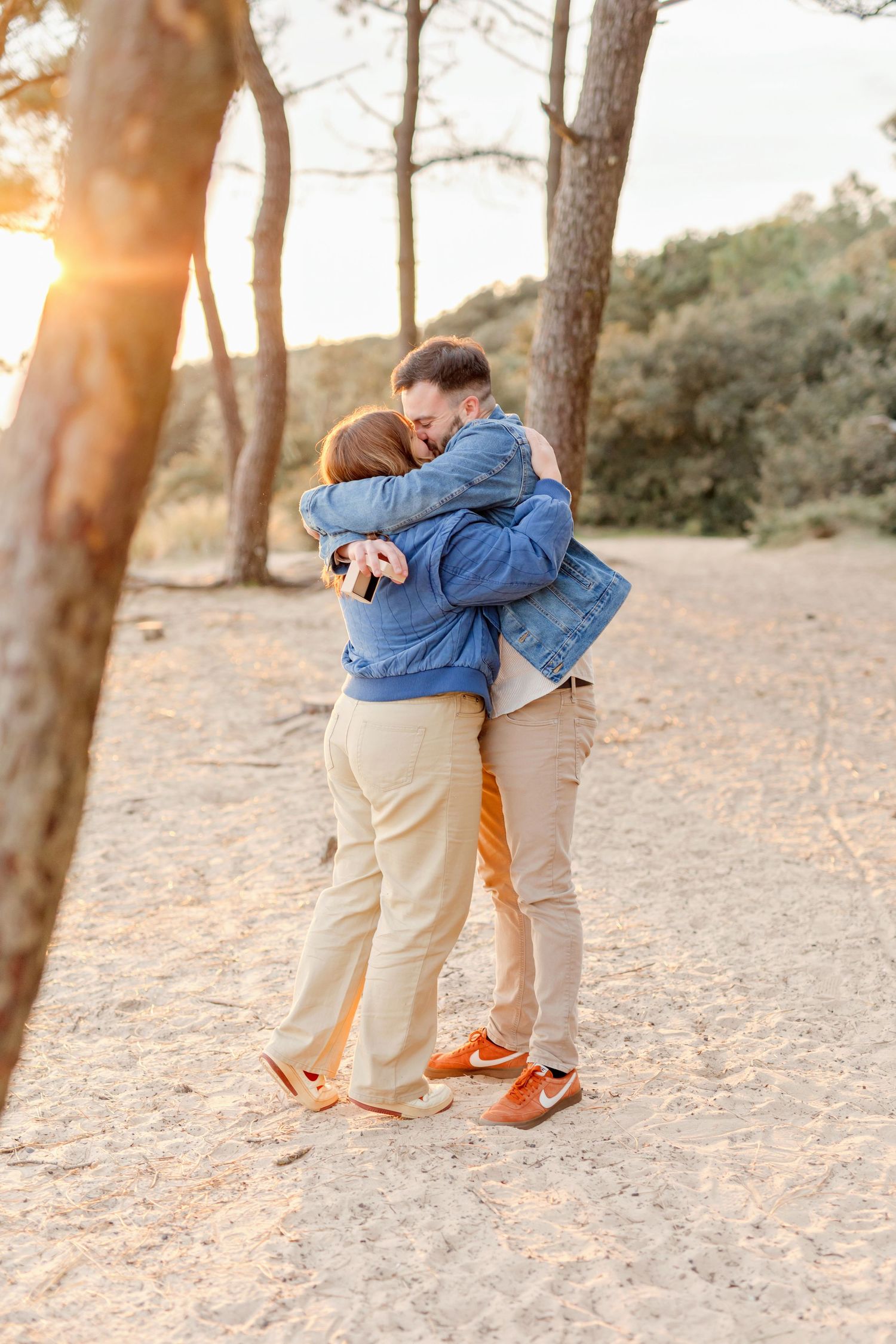 Two people in denim and khaki embrace joyfully with sunlight filtering through trees.
