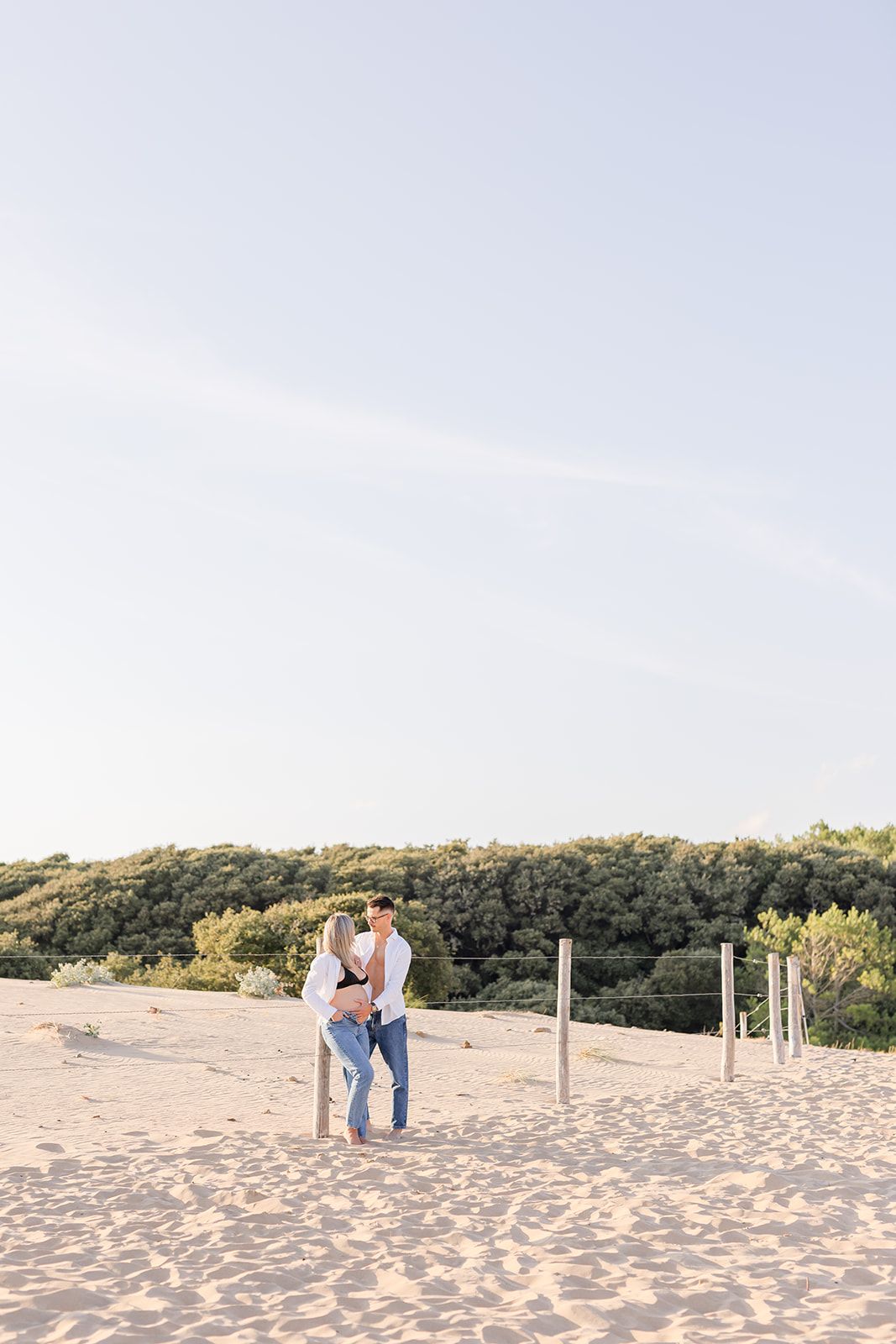 A couple stands together on a sandy beach shoreline with green shrubland in the background during golden hour.