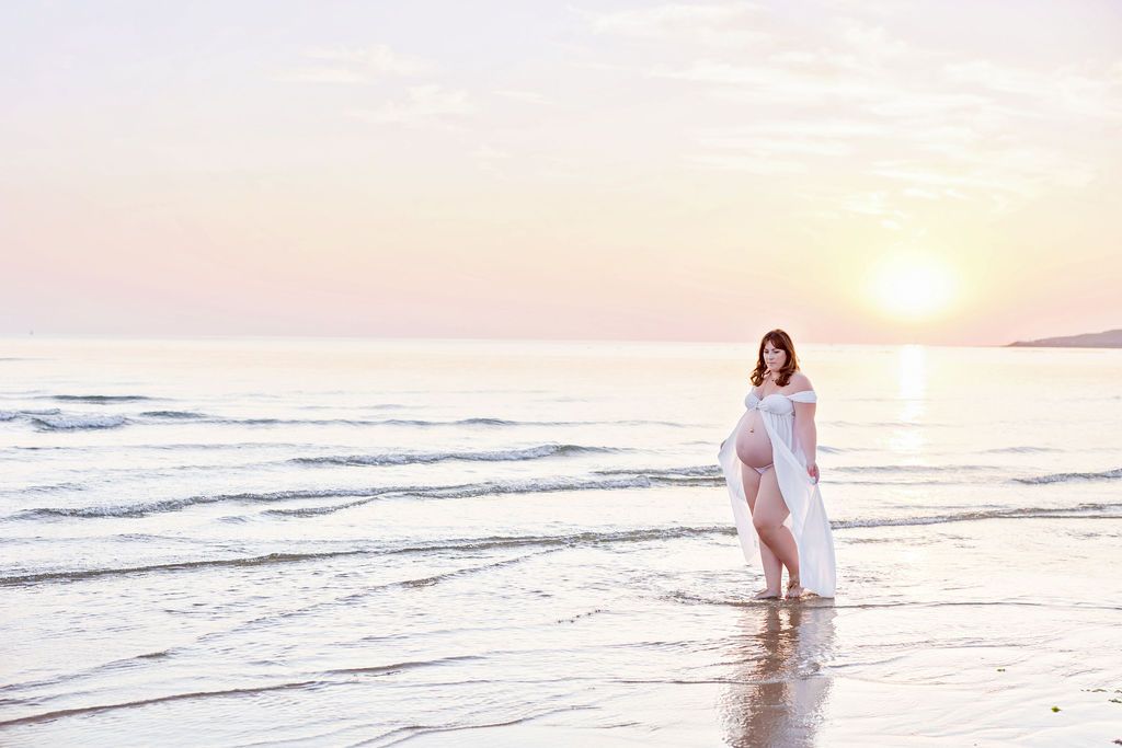 Figure in flowing white dress walks along peaceful beach shore during sunset.