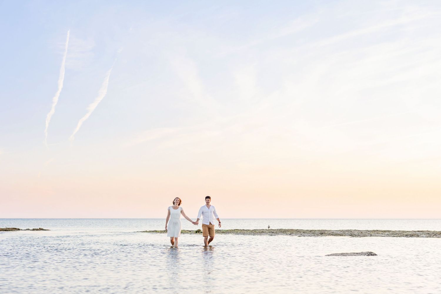 A romantic beach photoshoot captures a couple standing in shallow water during golden hour.