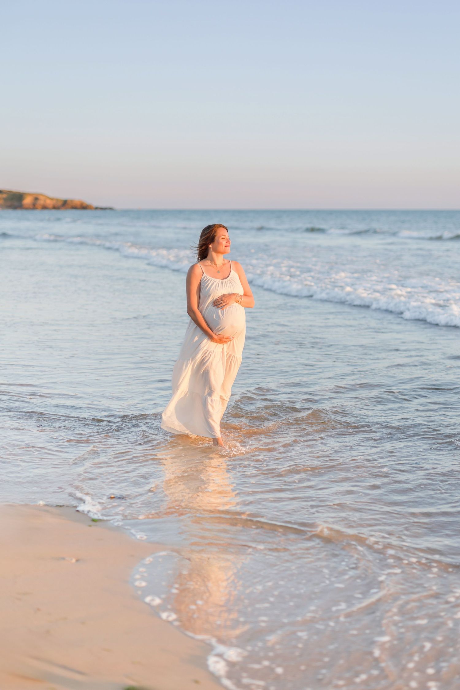 Expectant figure in white dress walking along beach shoreline at sunset.