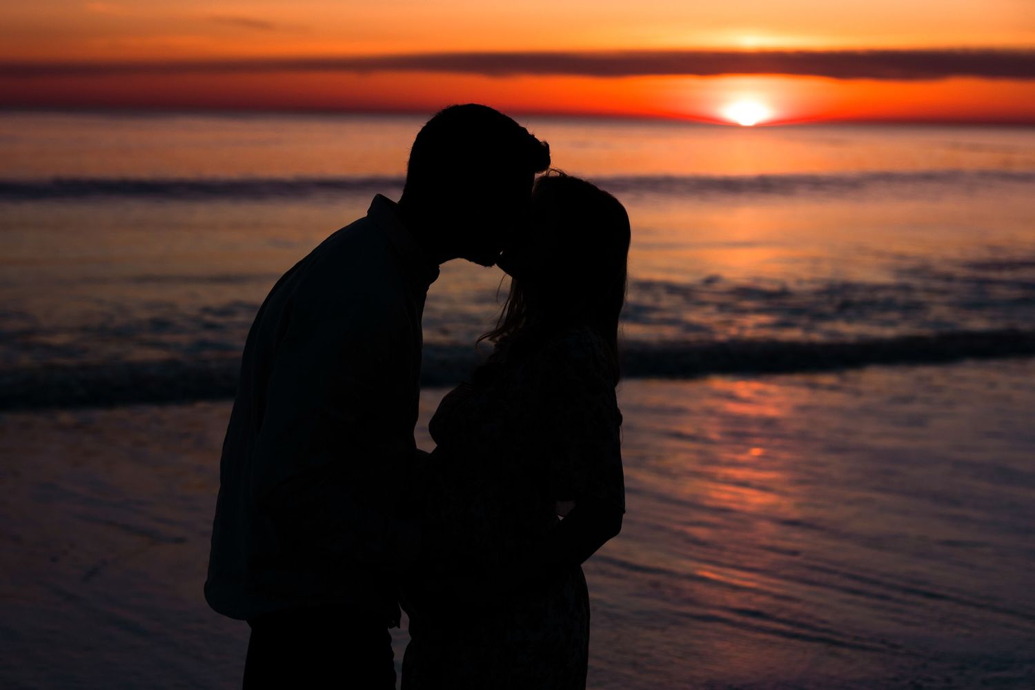 Romantic sunset silhouettes of couple by ocean with vibrant orange sky.