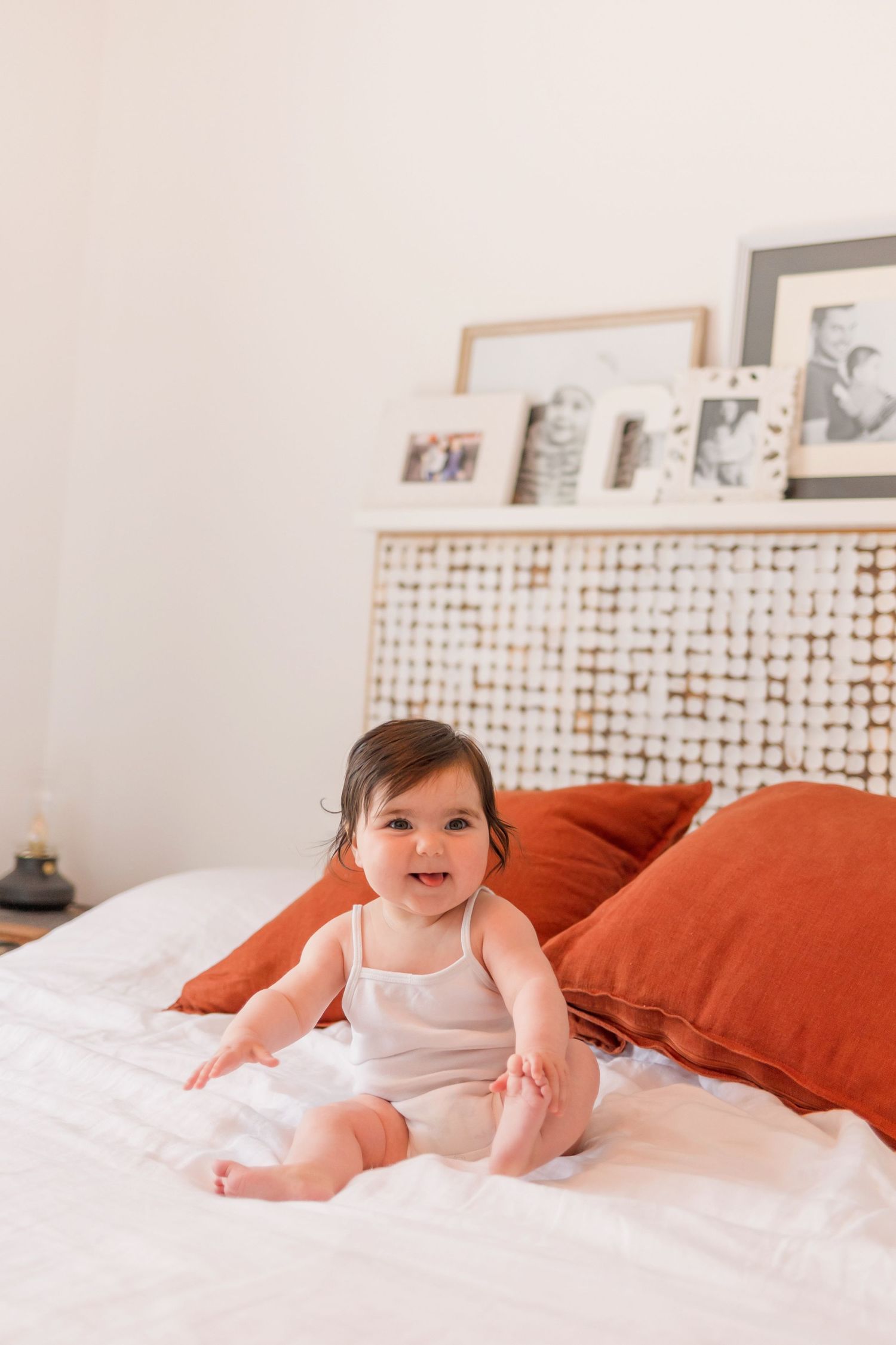 A baby in white sitting on a bed with orange pillows and photo frames on wall.