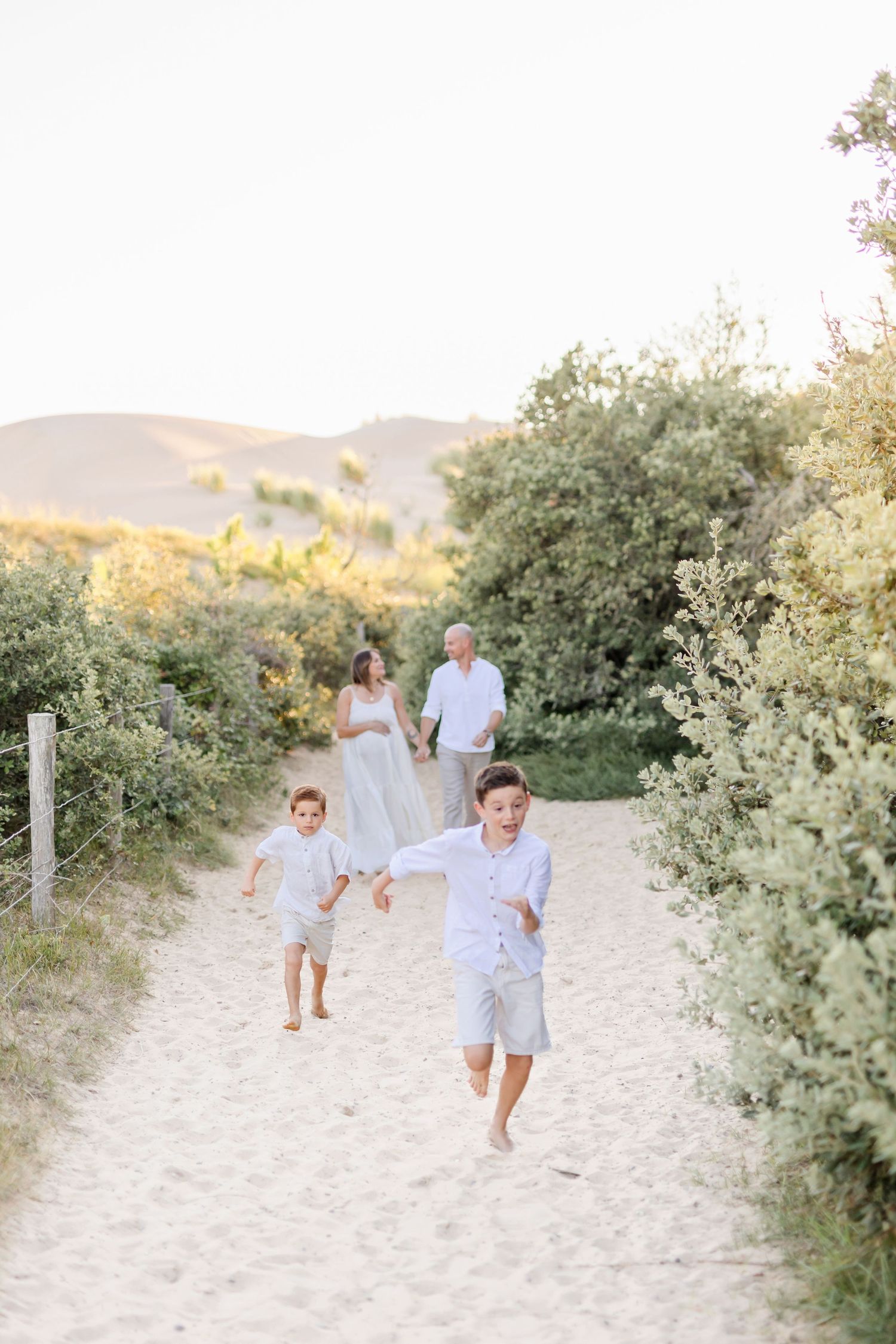 Family enjoying a playful moment on sandy path lined with coastal shrubs.