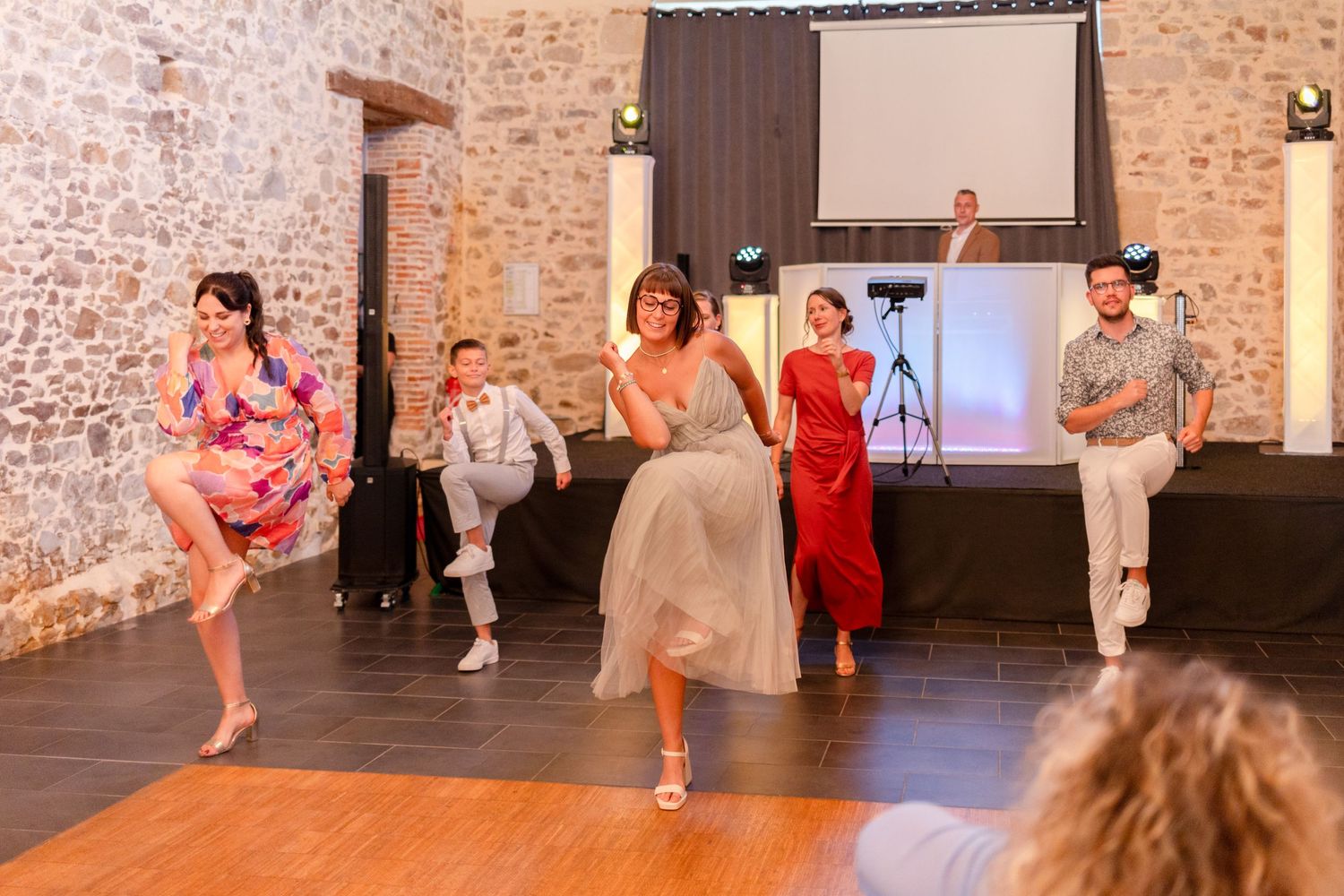 Group of wedding guests performing a choreographed dance routine on a dark wooden dance floor.
