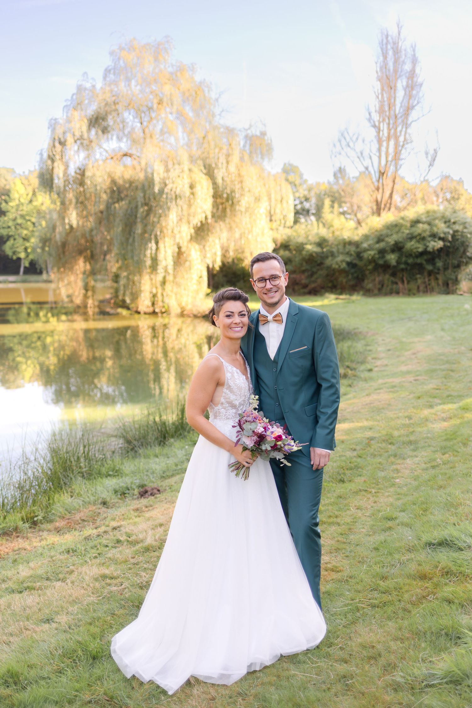 A wedding couple poses by a scenic pond with a weeping willow tree on a sunny day.