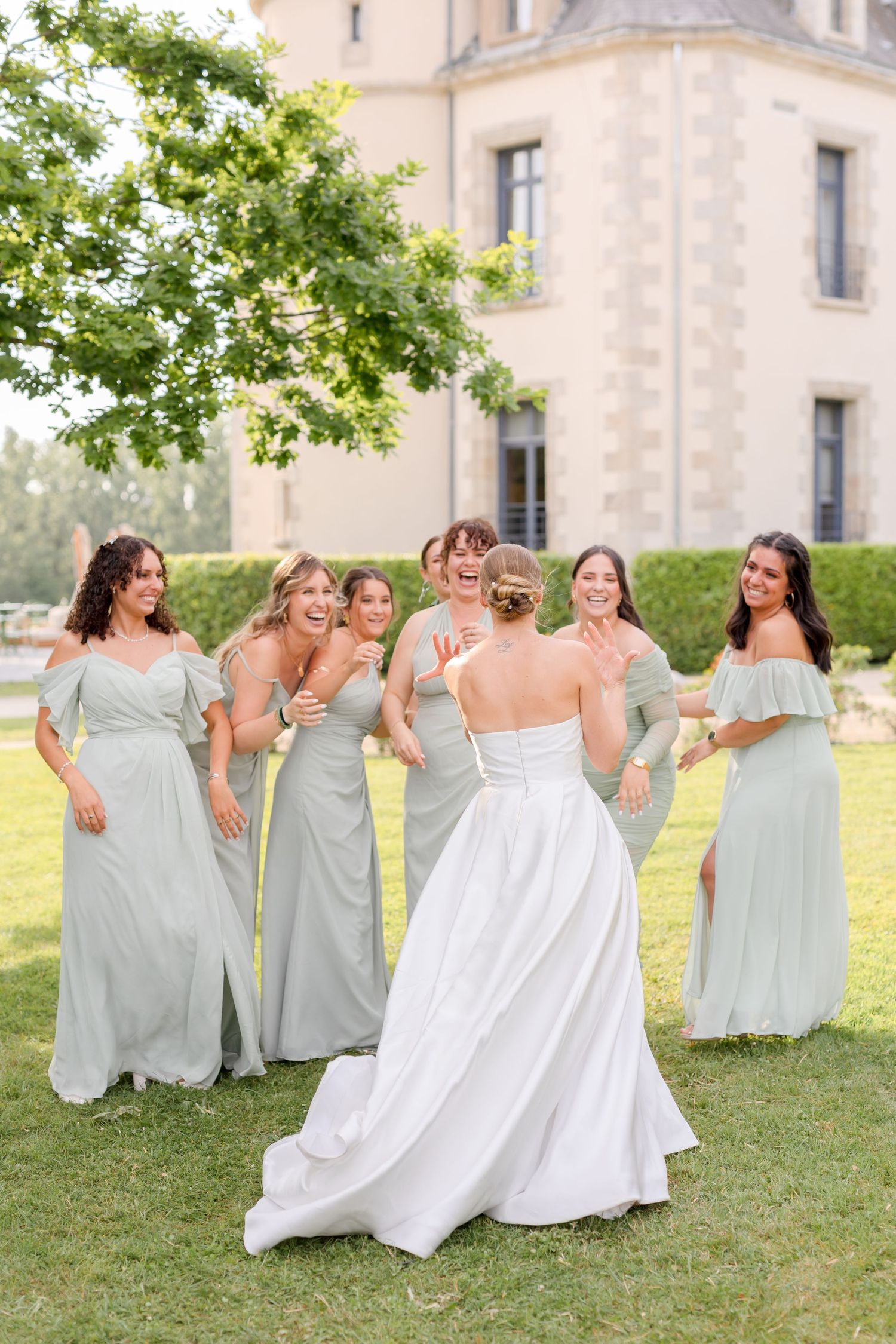 Wedding group strikes playful poses in matching mint dresses against historic chateau backdrop.