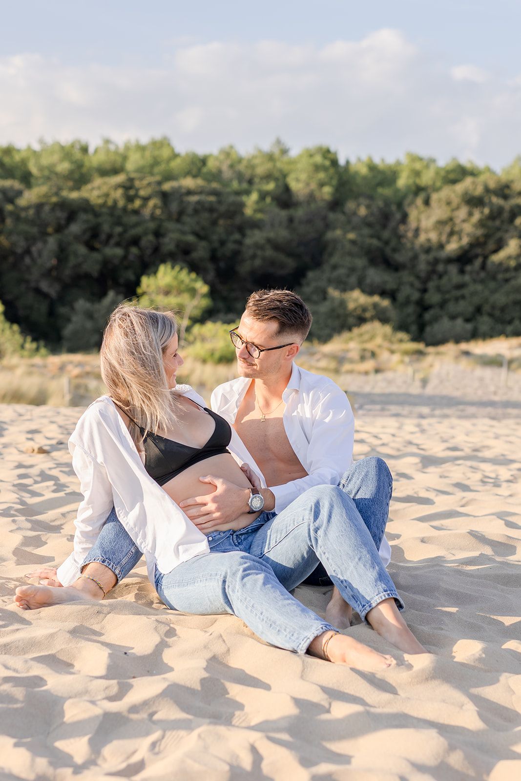 A romantic couple in casual attire lounges together on a sandy beach during golden hour.