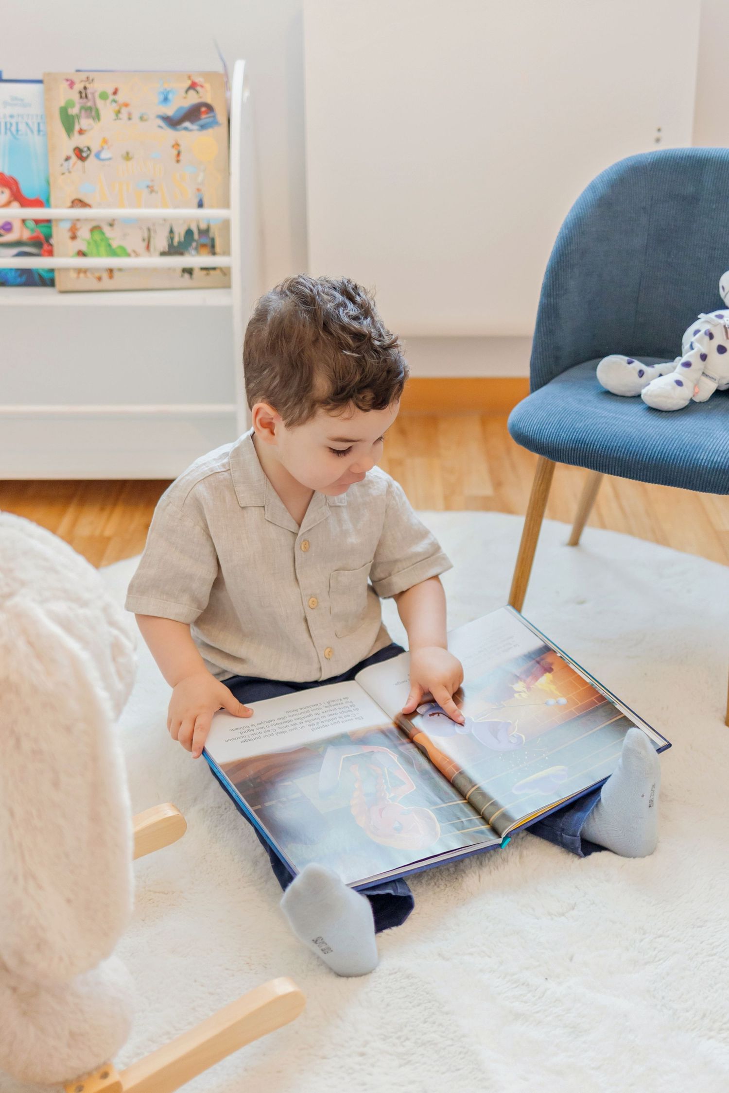 Young child sitting on floor reading a picture book in a modern room setting.