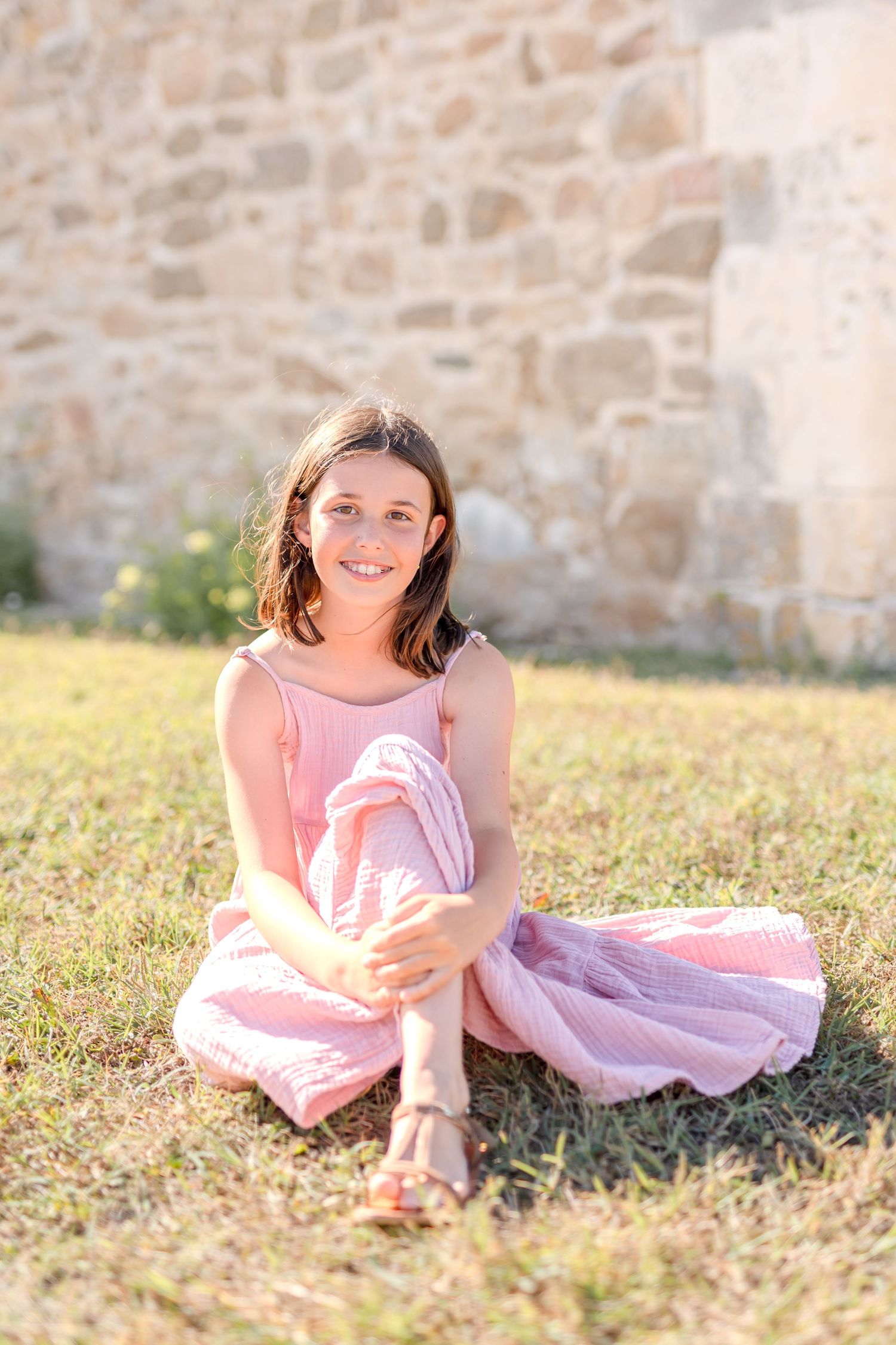 Young person in pink dress lying on grass near stone wall during golden hour.