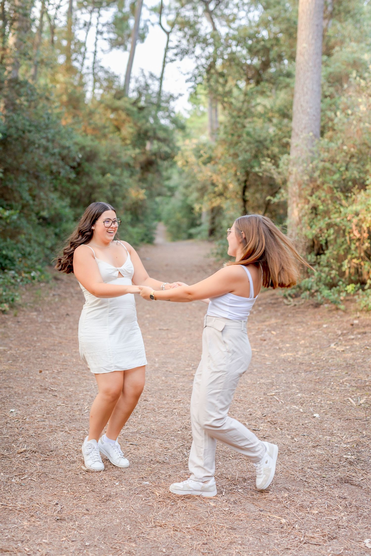 Two people in white outfits dancing and holding hands on a forest trail.