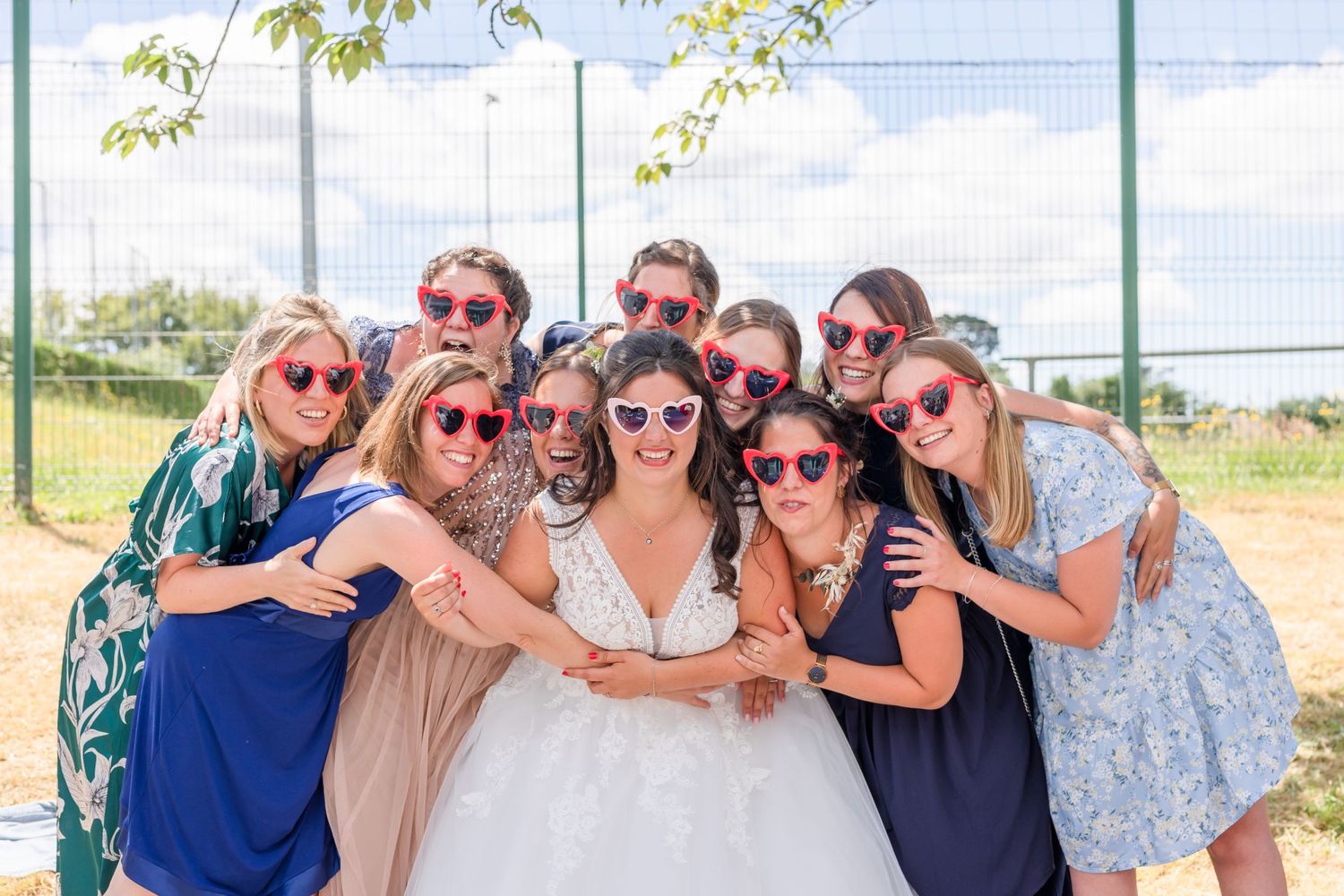 Group of friends in sunglasses pose together with bride at sunny outdoor wedding.