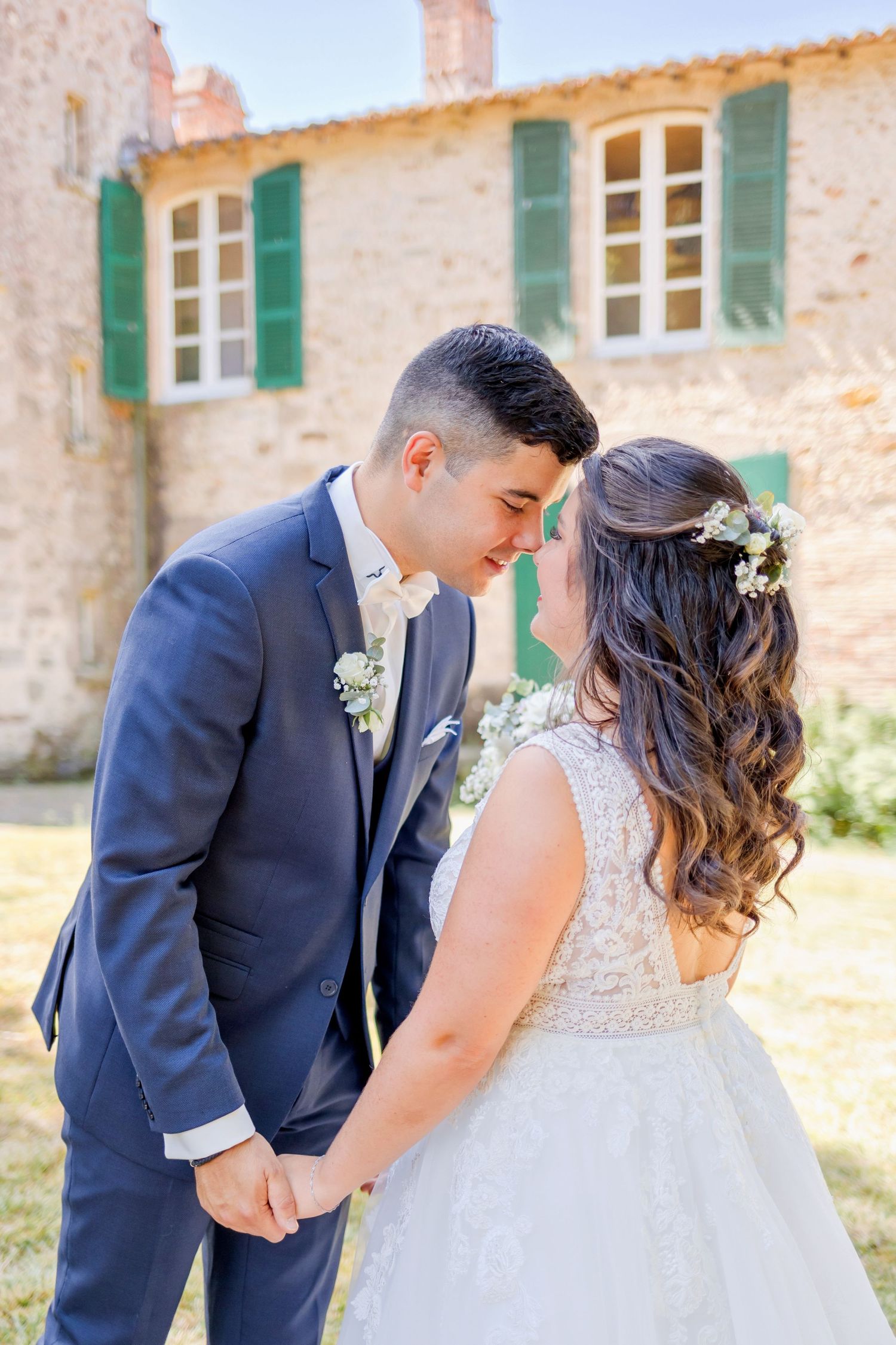 Elegant bride in lace gown and groom in navy suit embrace outside stone villa.