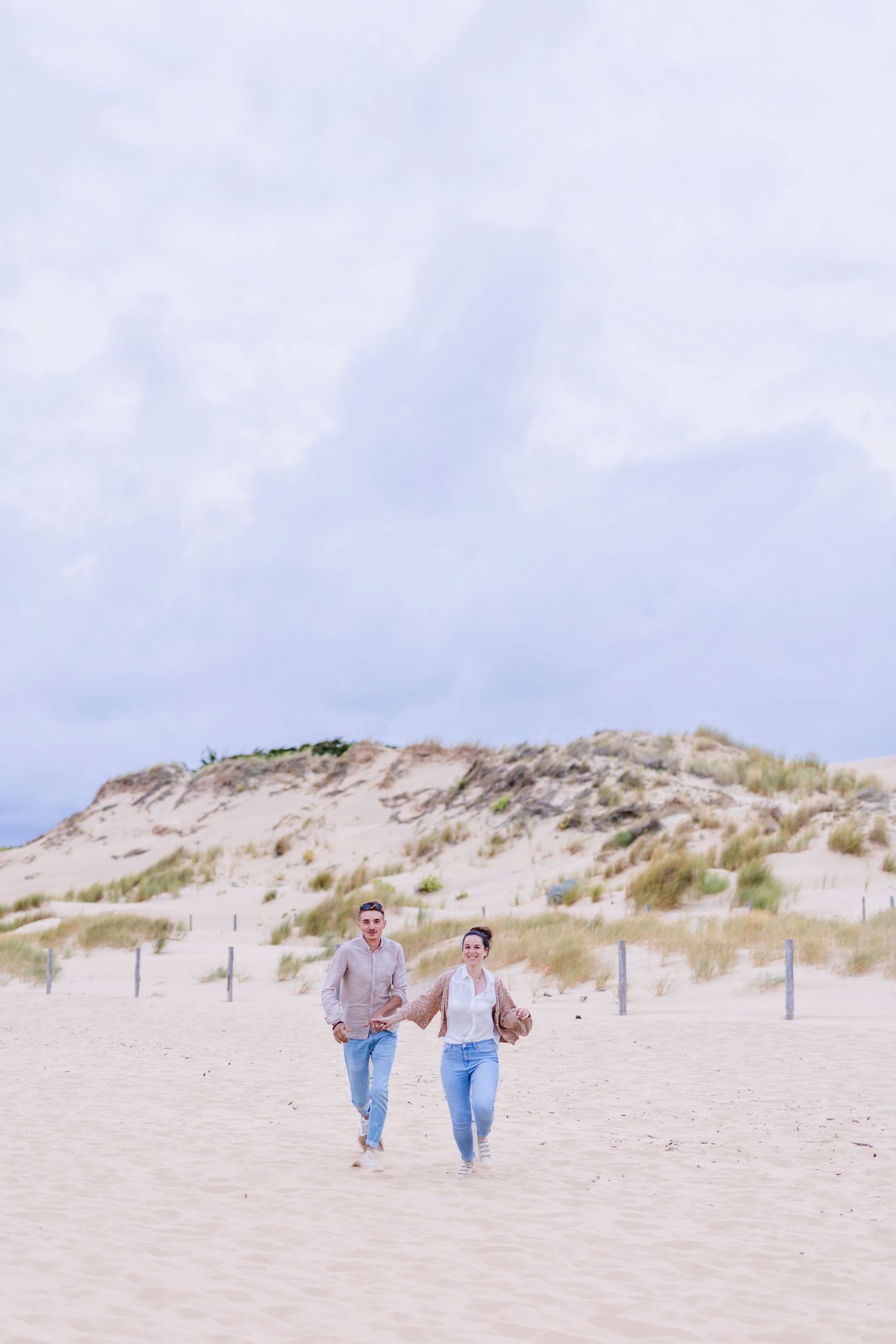 Couple running together on beach with sand dunes and blue sky in background.