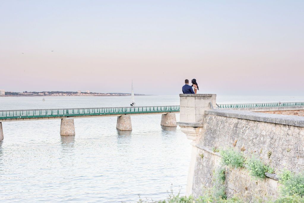 A coastal stone wall and bridge with pastel sunset colors reflecting on calm waters.