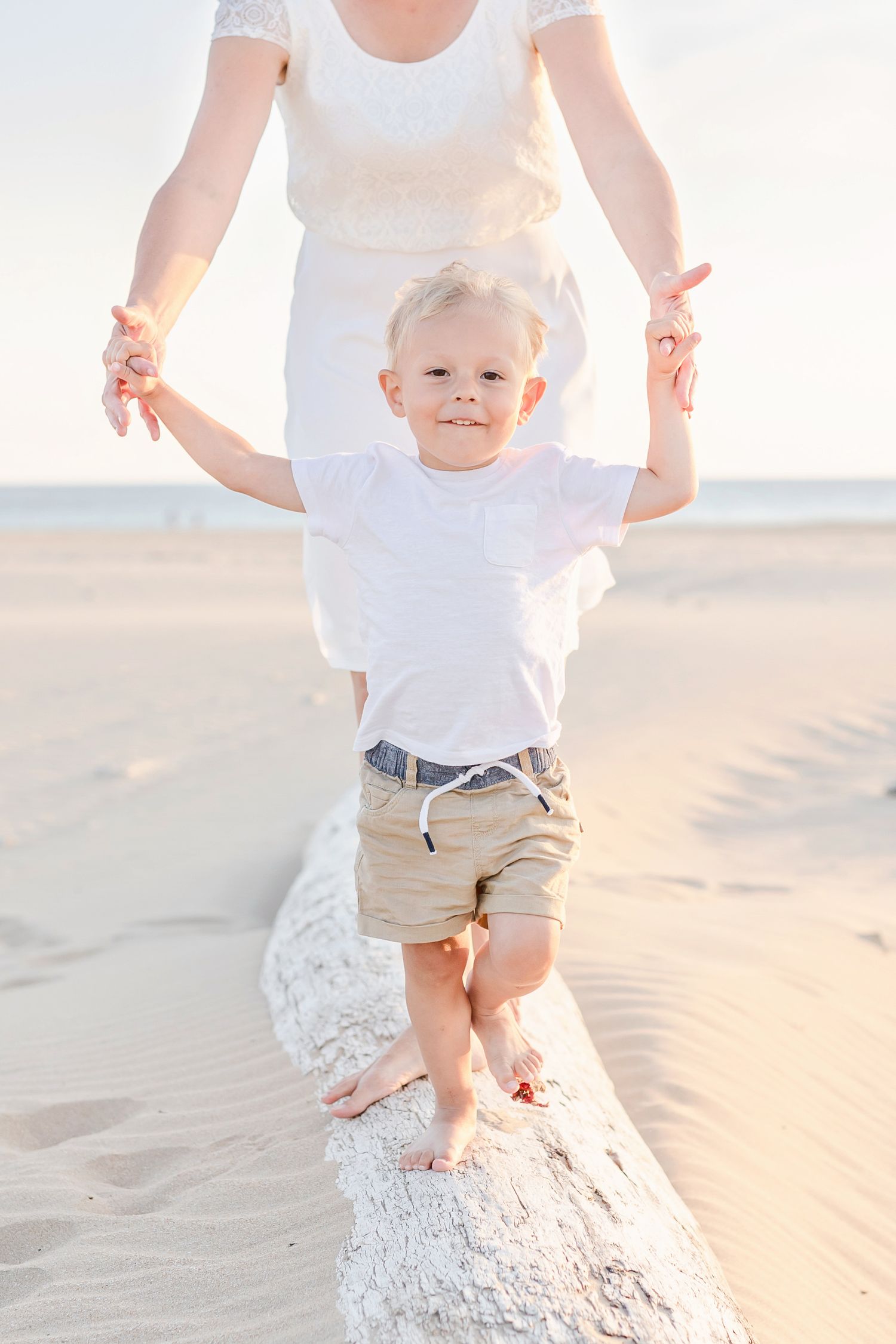 A toddler in white shirt and khaki shorts balances on a sand ridge at the beach while holding hands with someone.