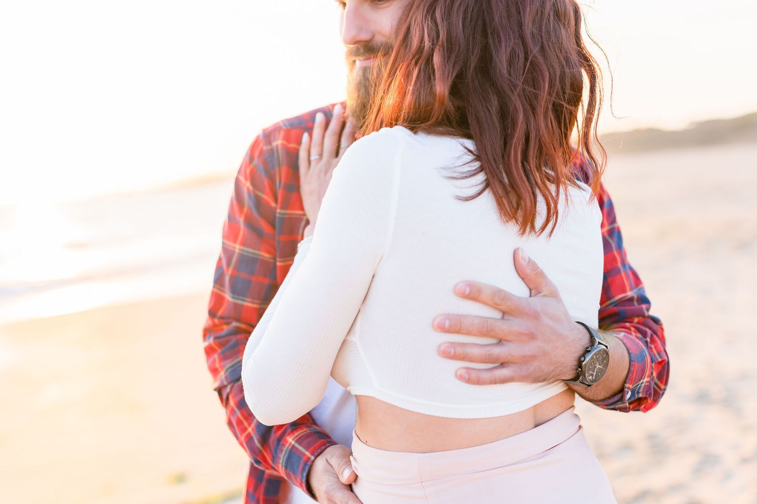 A pregnant woman in a white dress and plaid shirt poses on a sandy beach during sunset.