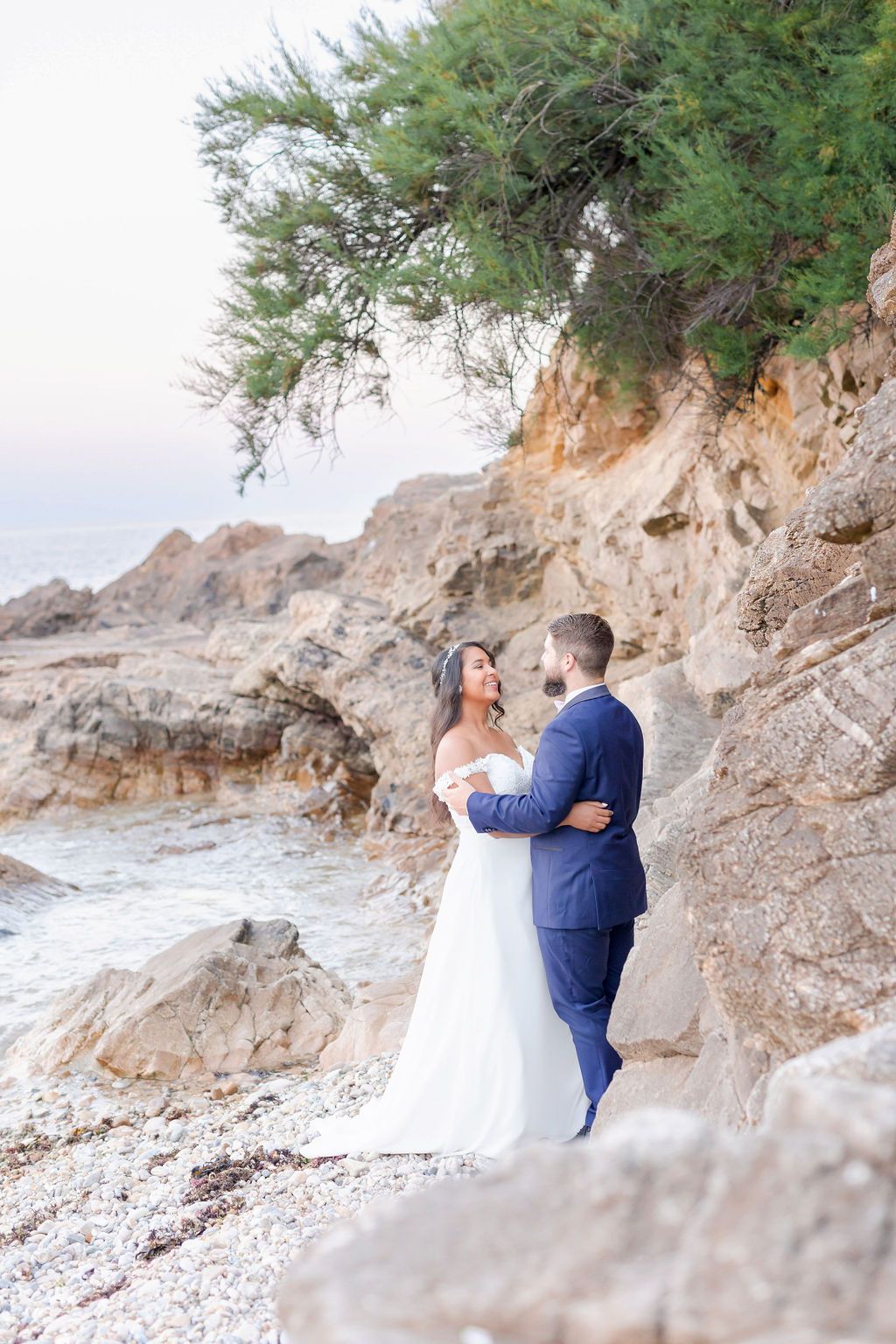 A couple in wedding attire shares an intimate moment on a rocky coastline with waves and cliffs in the background.