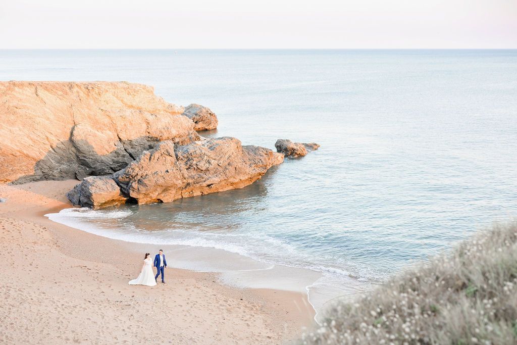 A couple in wedding attire strolls on a secluded beach surrounded by rugged coastal cliffs.