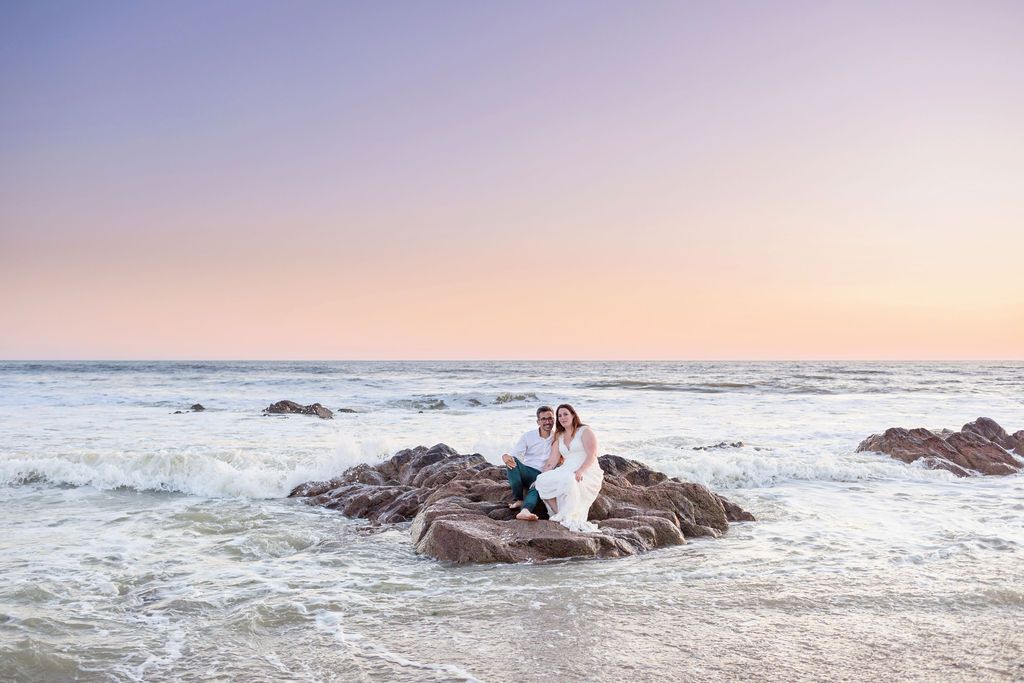 A couple poses on rocky beach outcrop during pastel sunset.