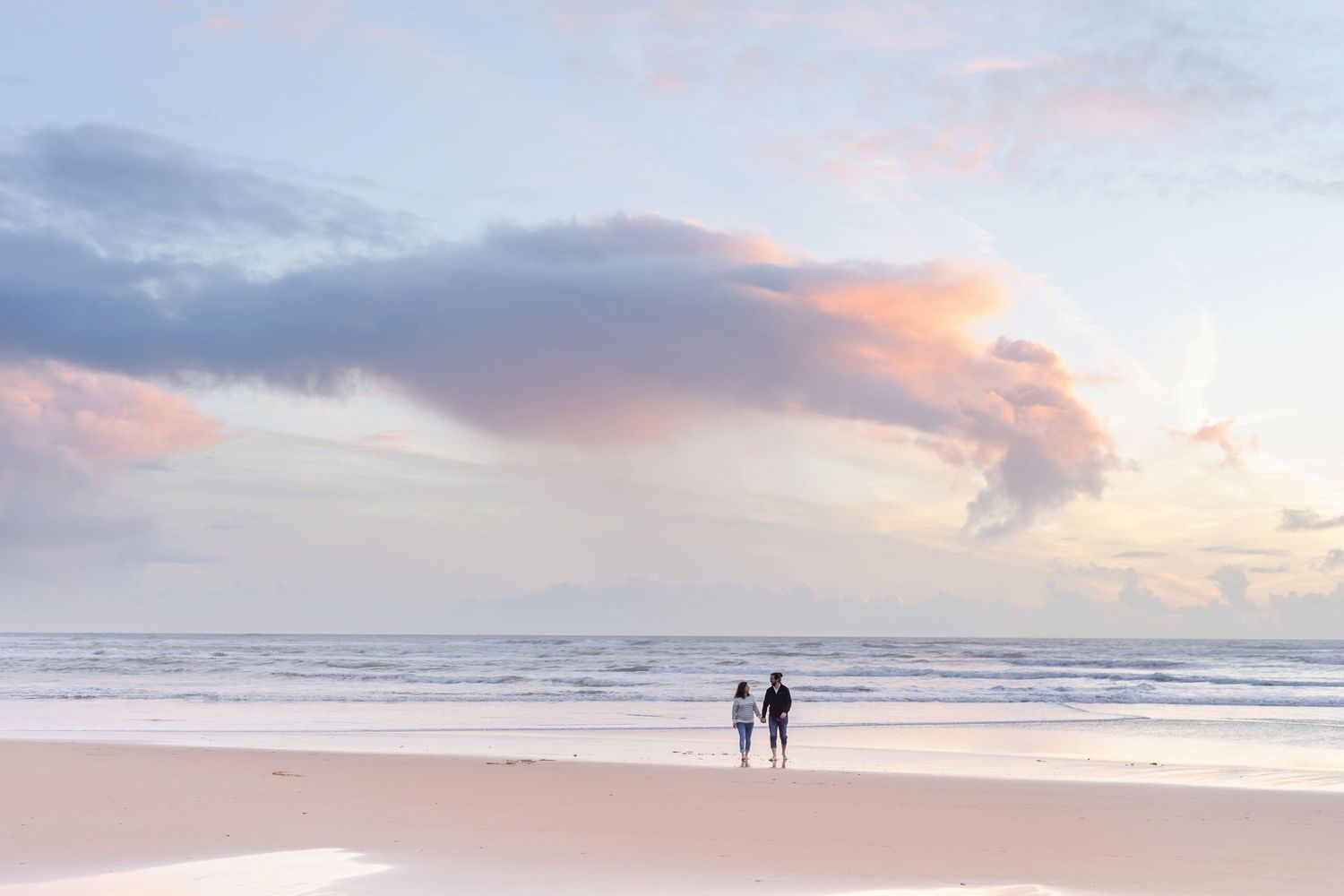 Two distant figures stand on an empty beach during a pastel sunset.