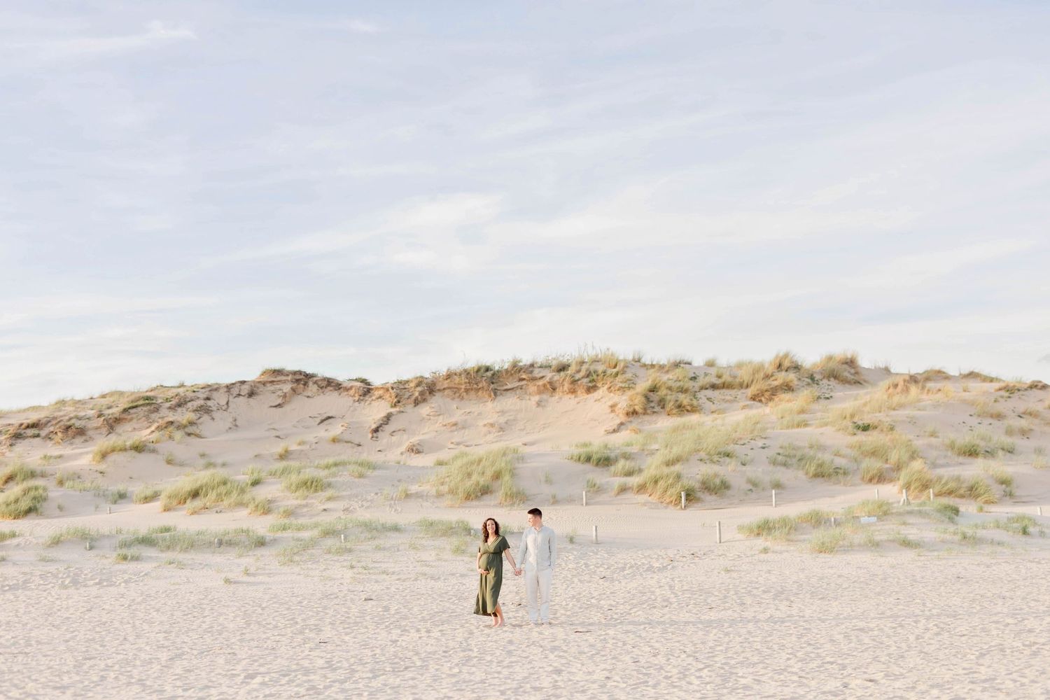 A distant couple stands together on sandy beach with dunes in background.