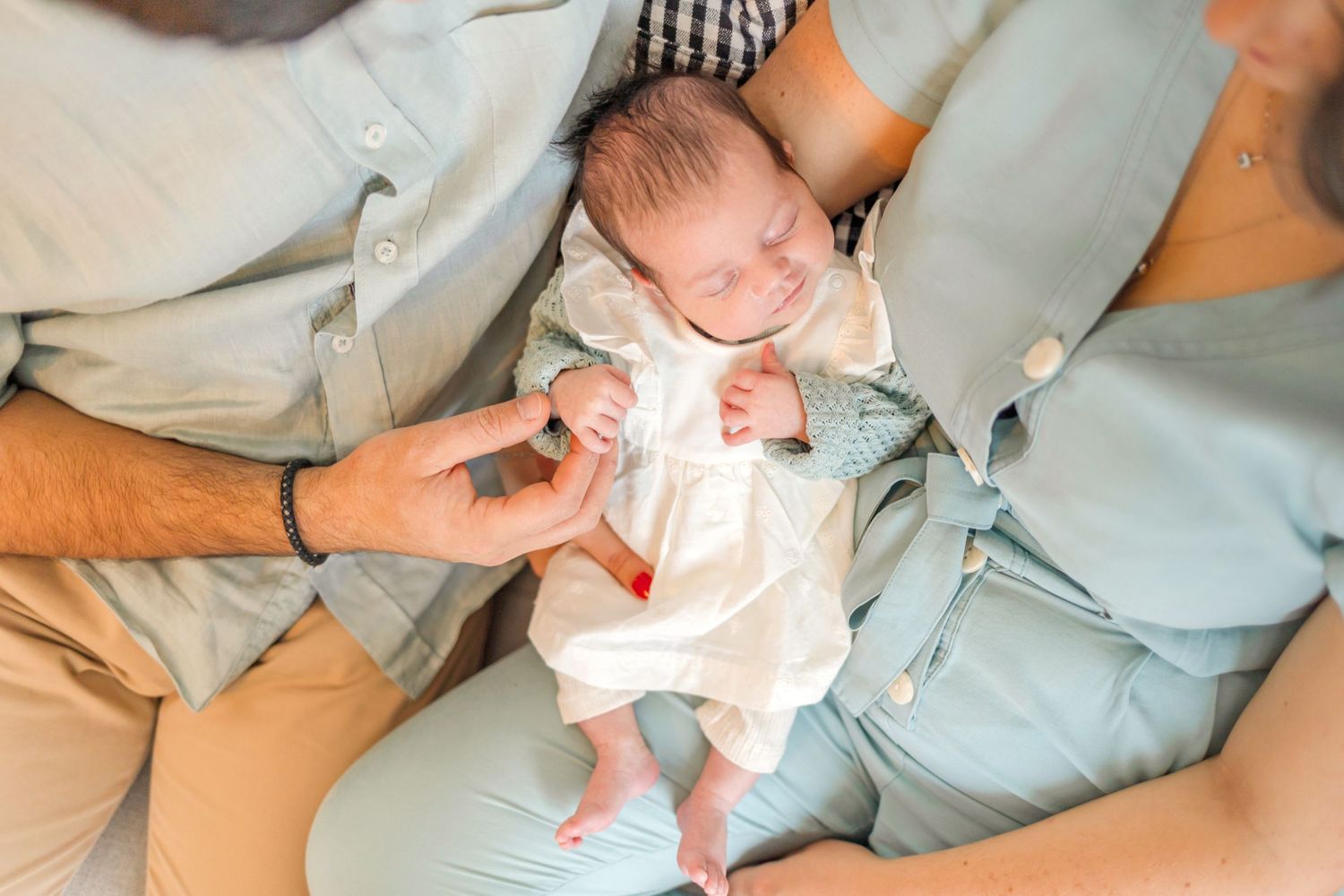 Newborn baby in white outfit being cradled between parents on couch.