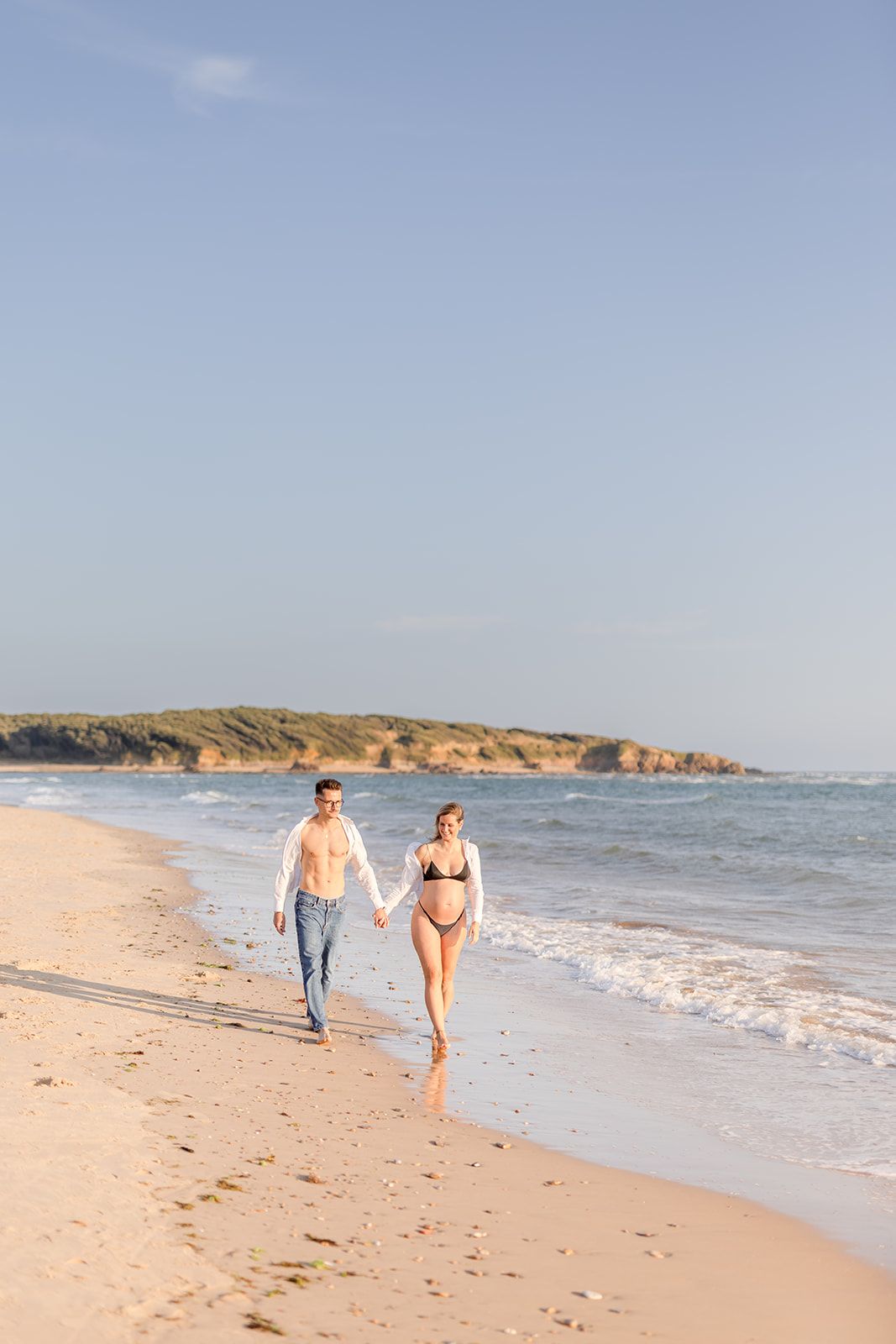 Two people walking together along a sandy beach shoreline with ocean waves and rocky cliffs in the background.