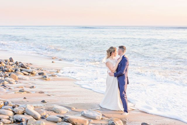 Wedding couple walking along rocky beach shoreline during sunset.