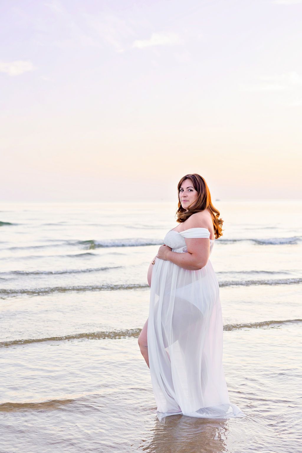 Expecting mother in flowing white dress poses on beach during sunset.