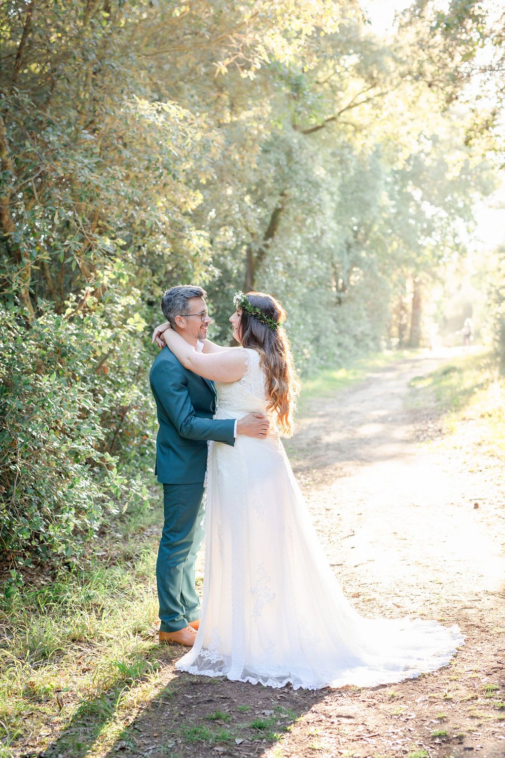 Newlyweds share a tender moment on a sunlit forest path during their wedding photoshoot.