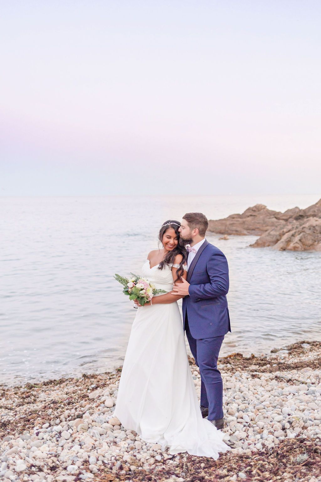 Joyful wedding moment captured on rocky beach with soft pink sunset lighting.
