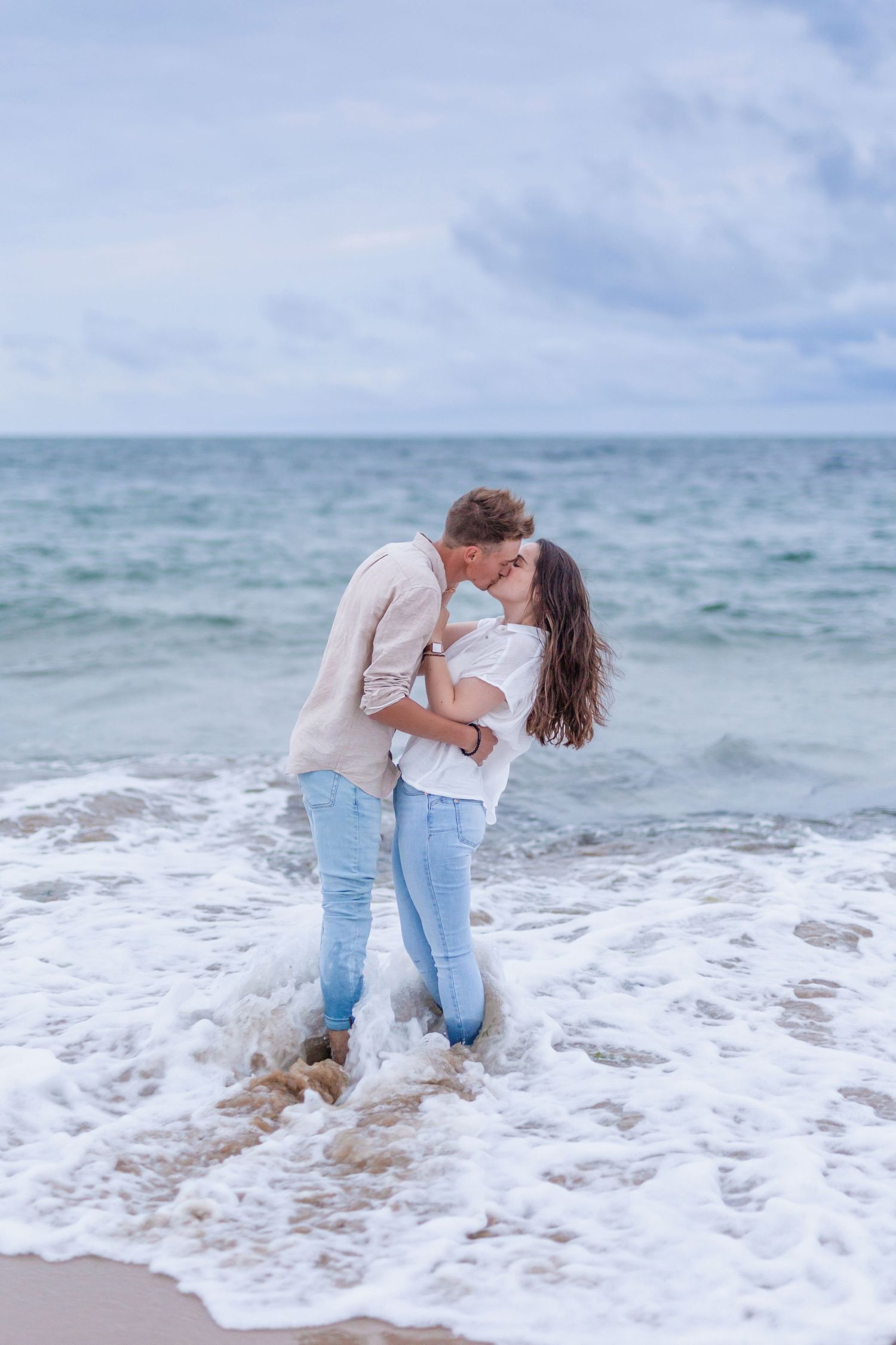 A romantic couple embraces while standing in ocean waves on a cloudy beach day.